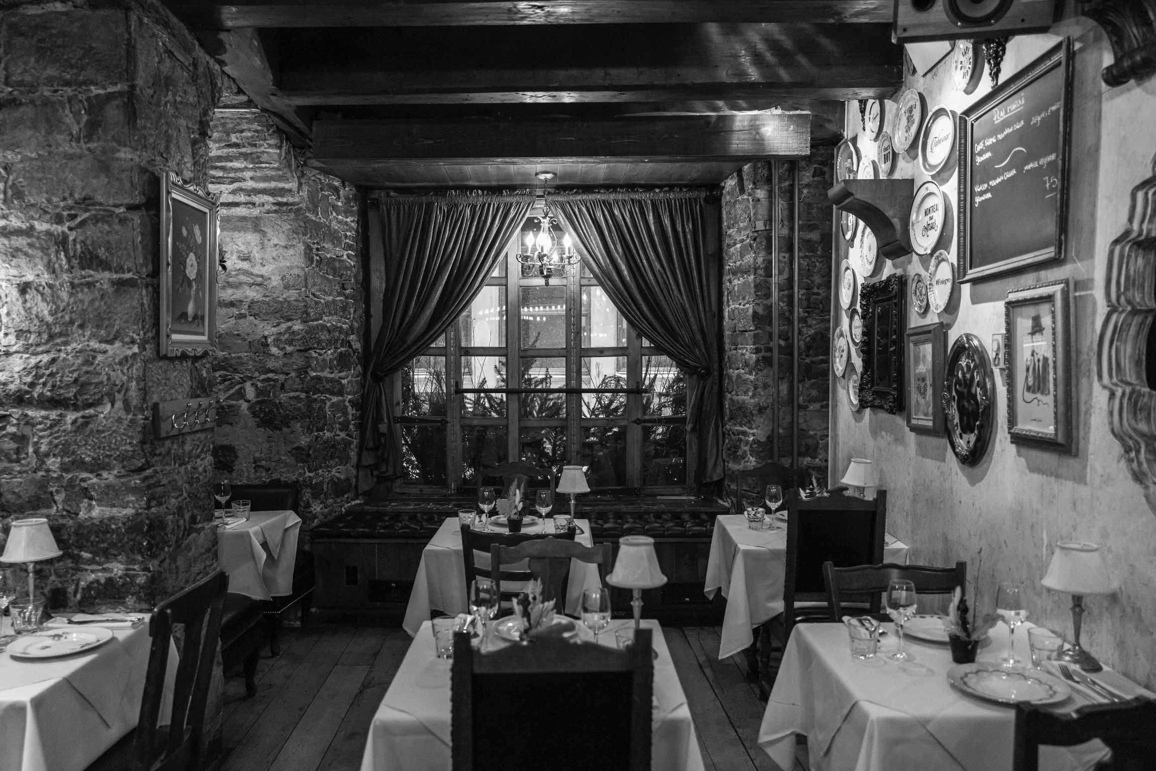 Une salle de restaurant élégante avec des murs en pierre, des tables avec nappes blanches, des verres à vin et des lampes de table, et une grande fenêtre avec des rideaux en velours, éclairée par un lustre suspendu.