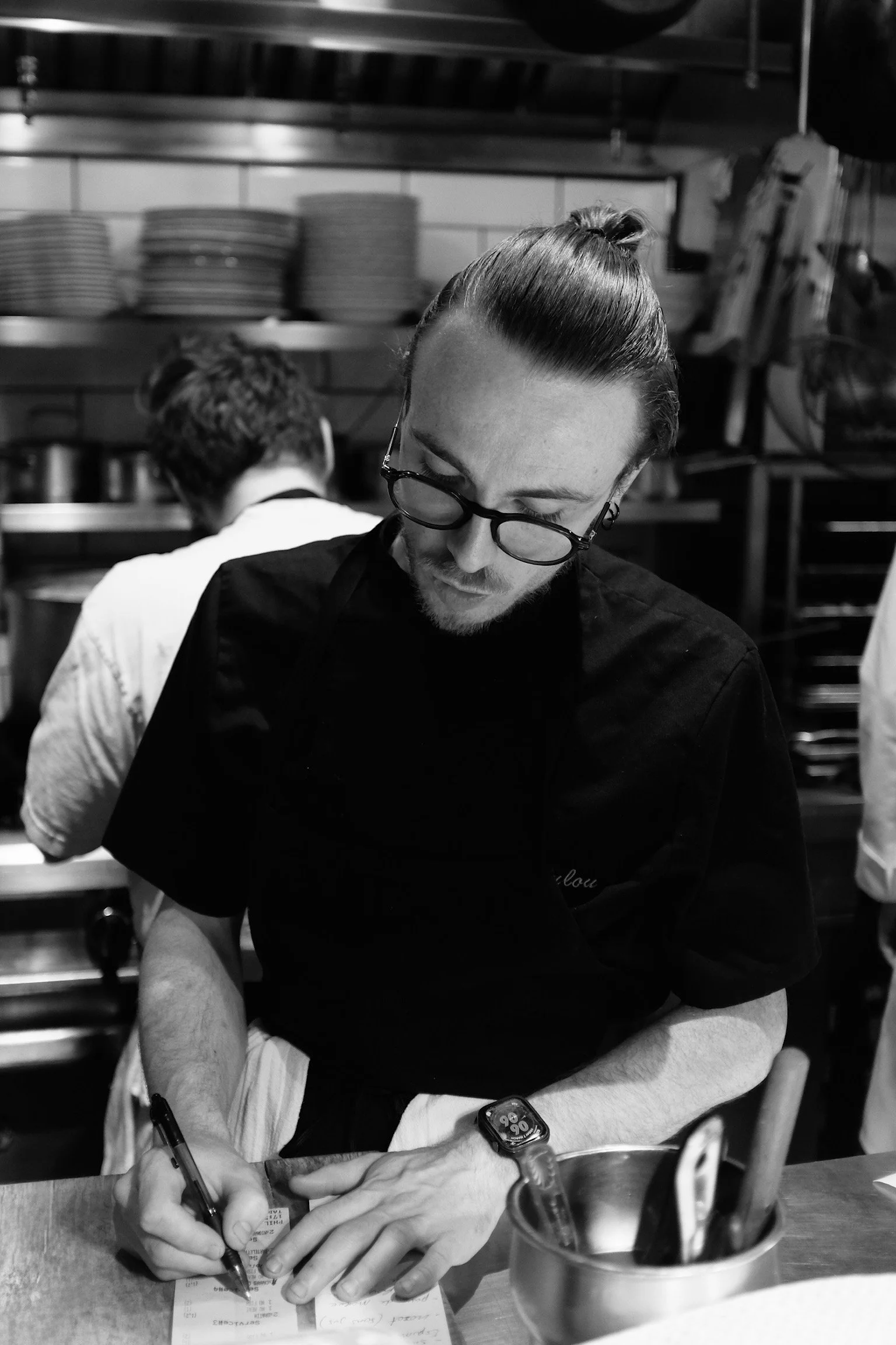 Un chef ou cuisinier écrit quelque chose sur un menu ou une commande dans une cuisine professionnelle, portant des lunettes et un bracelet connecté.