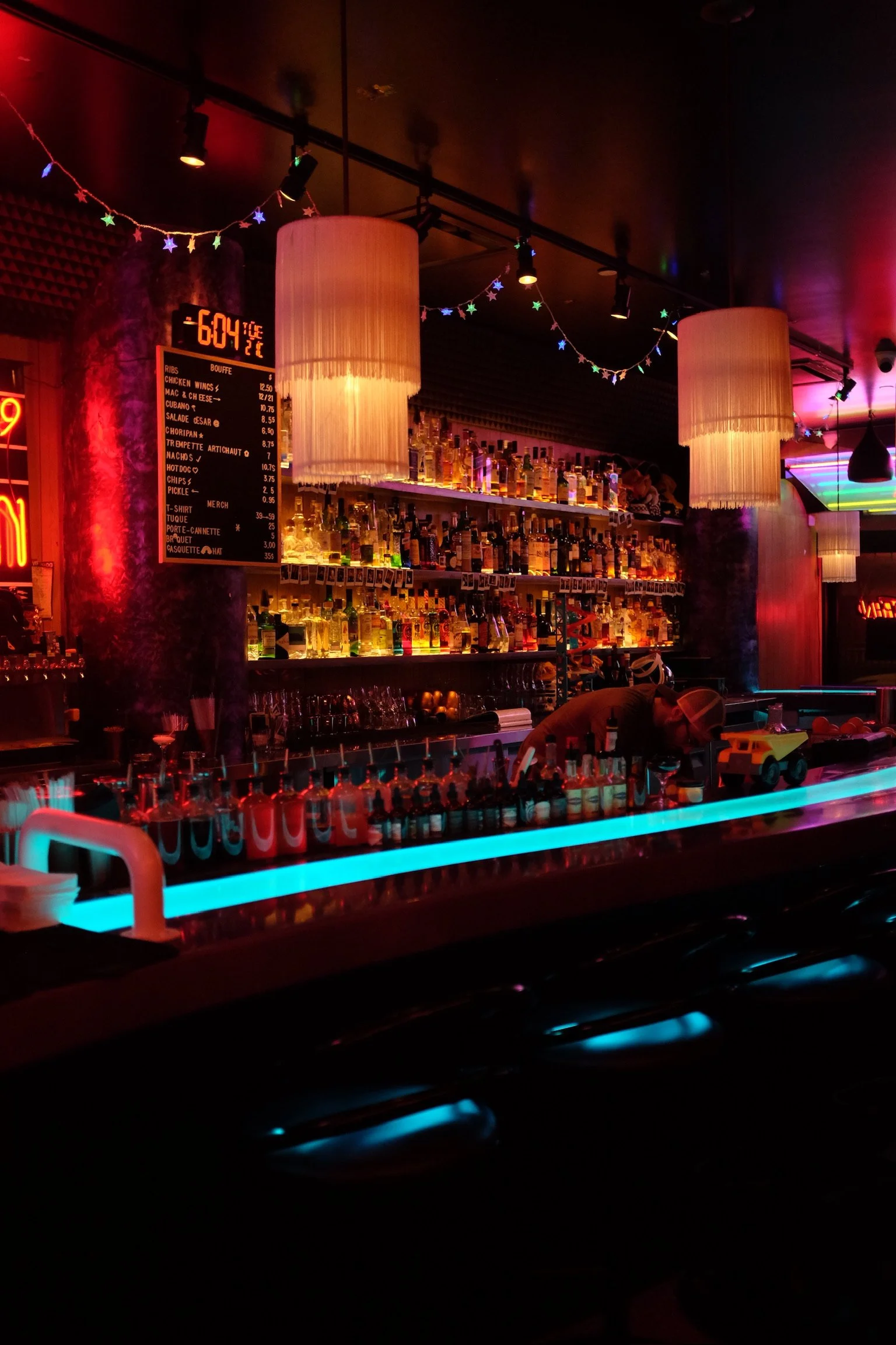 Un bar avec une étagère remplie de bouteilles d'alcool illuminées, un tableau avec le menu, lumières colorées, et un barman en train de préparer des drinks dans une ambiance sombre et colorée.