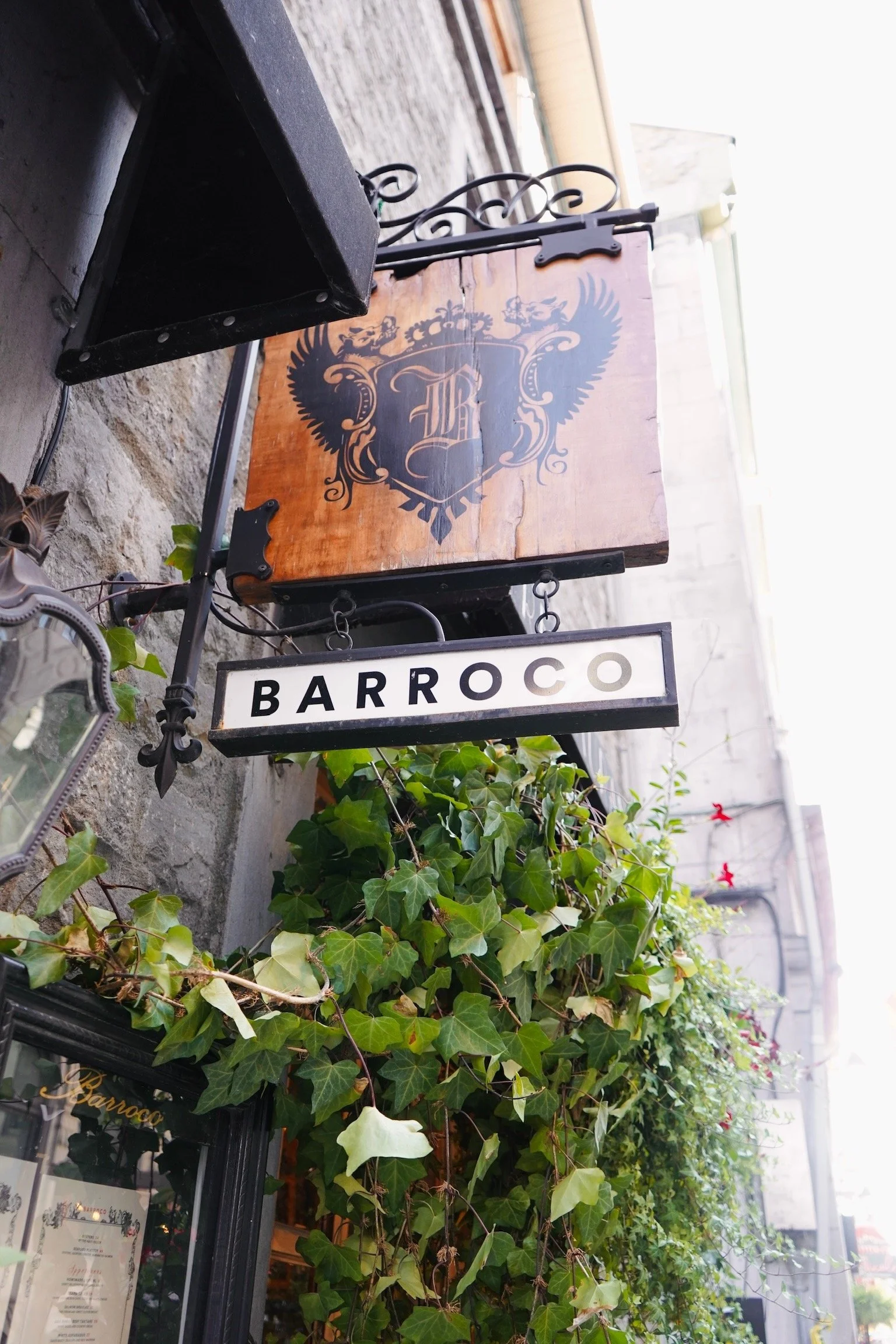Panneau de l'établissement "Barroco" avec un symbole orné d'ailes et de lettres gothiques, entouré de plantes vertes grimpantes, dans une rue commerçante.