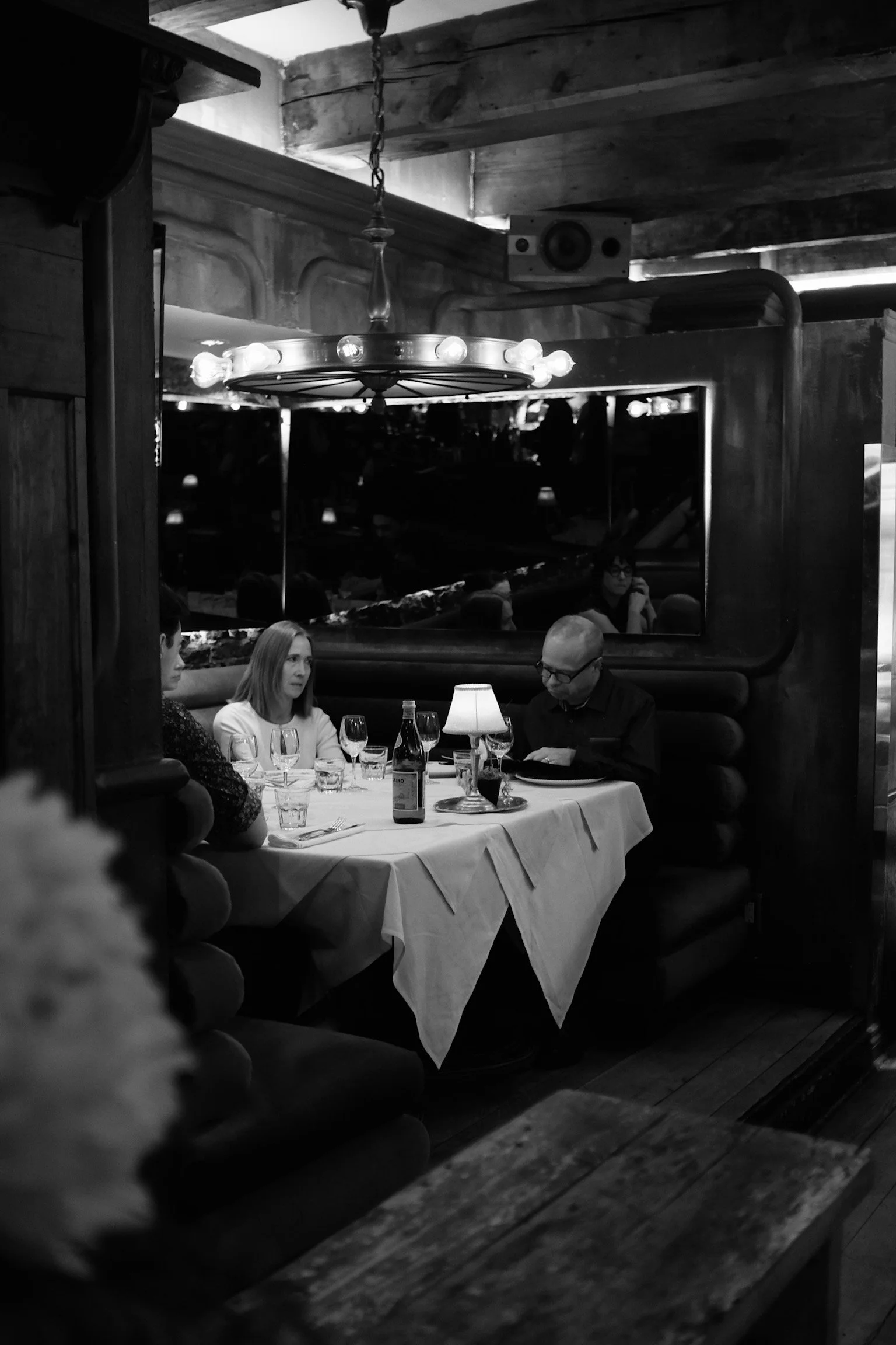 Une scène de restaurant avec trois personnes assises à une table, décor en bois, lampes, bouteilles et verres sur la table, reflet dans le miroir et ambiance tamisée.