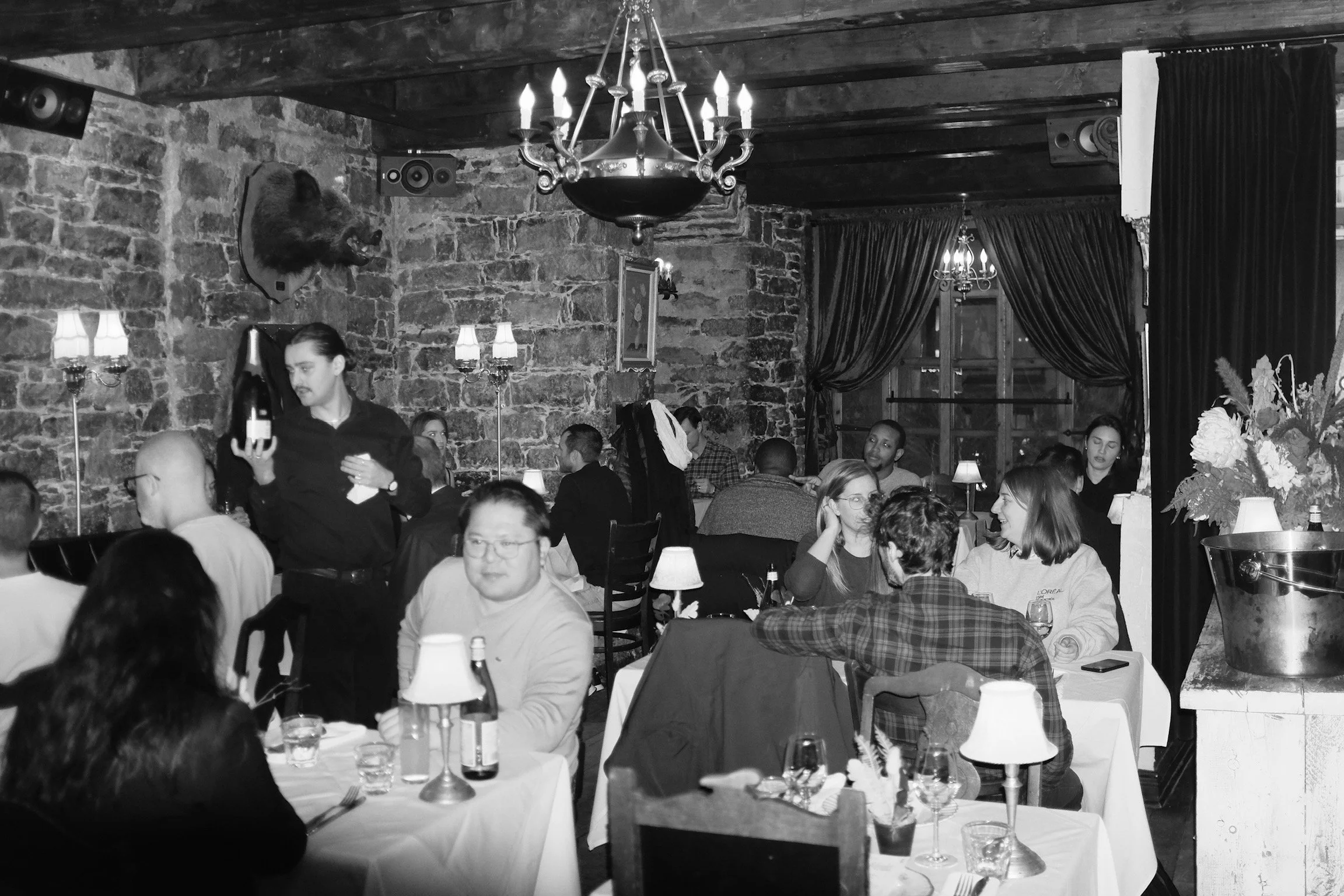 Recueil d'images d'une salle à manger de restaurant avec des clients assis à des tables avec des lampes, un serveur servant du vin et décor rustique avec des murs en pierre et des rideaux, en noir et blanc.