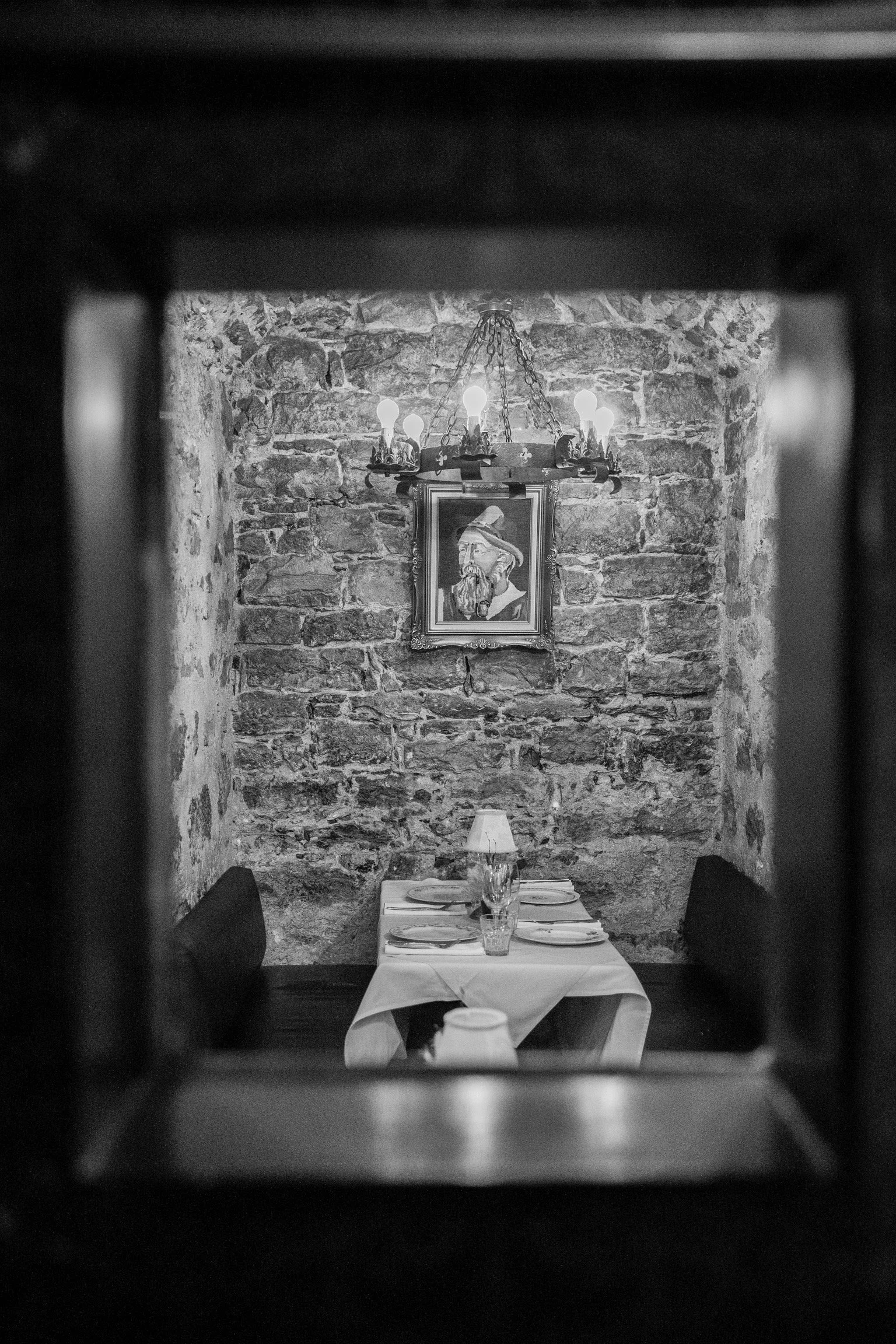 Salle à manger avec table préparée pour un repas, mur en pierre, tableau en portrait et chandelier en métal.
