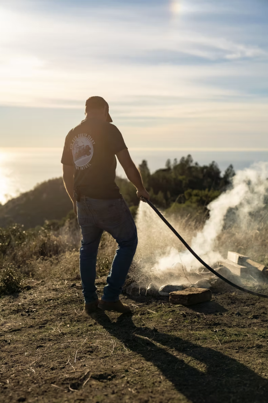 A man standing on a dirt road extinguishing a campfire with a water hose