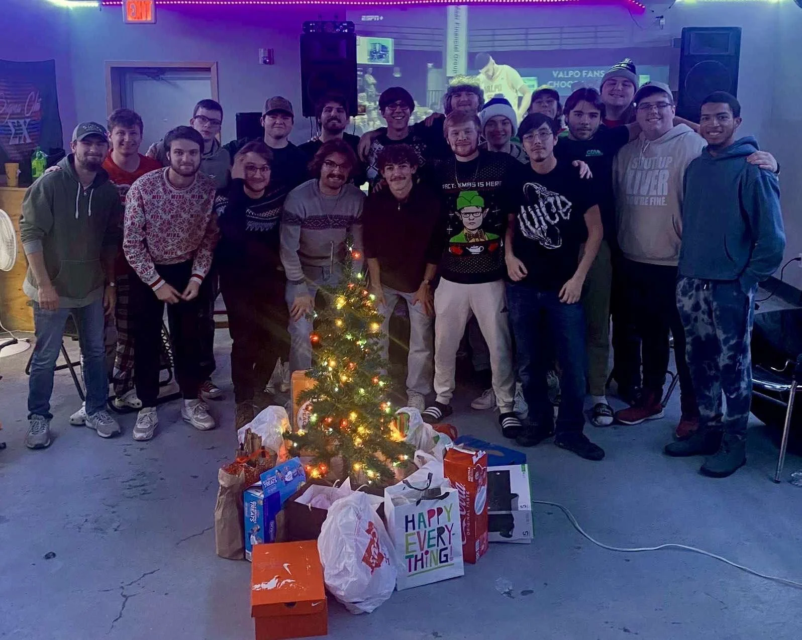 A group of young men gathered around a small decorated Christmas tree with wrapped gifts underneath, in an indoor setting with purple and blue lighting, celebrating the holidays.