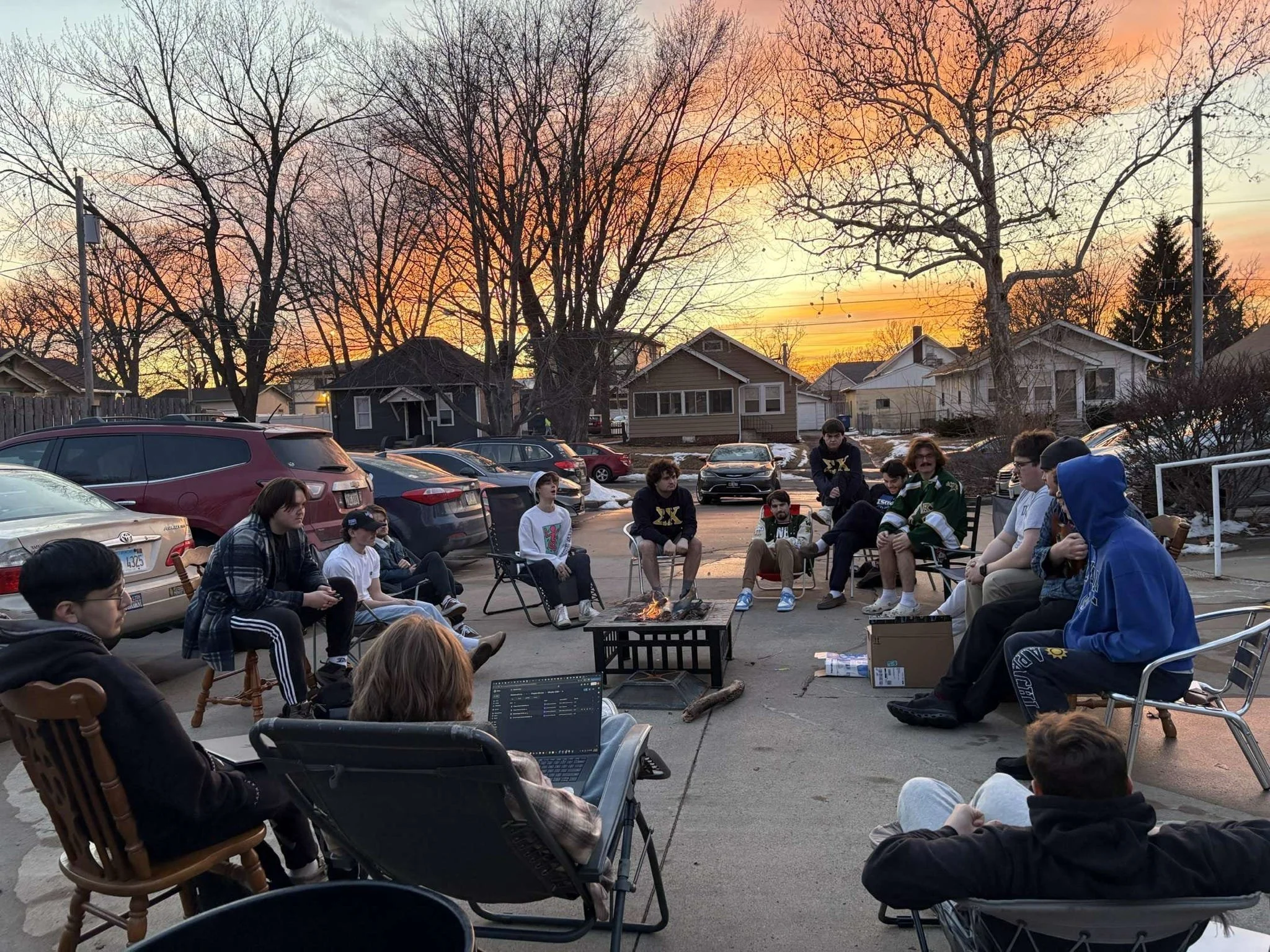 A group of people sitting around a fire pit outdoors during sunset, in a residential neighborhood, enjoying a gathering.