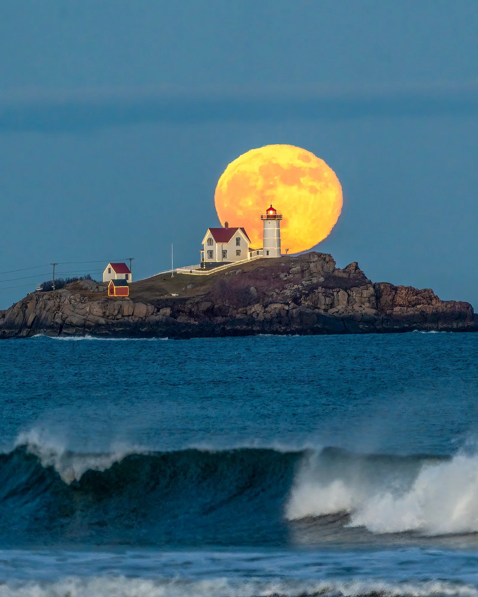 Bright yellow full moon rising over white and red lighthouse and house at Nubble Beach, Maine, with ocean wave in the foreground.