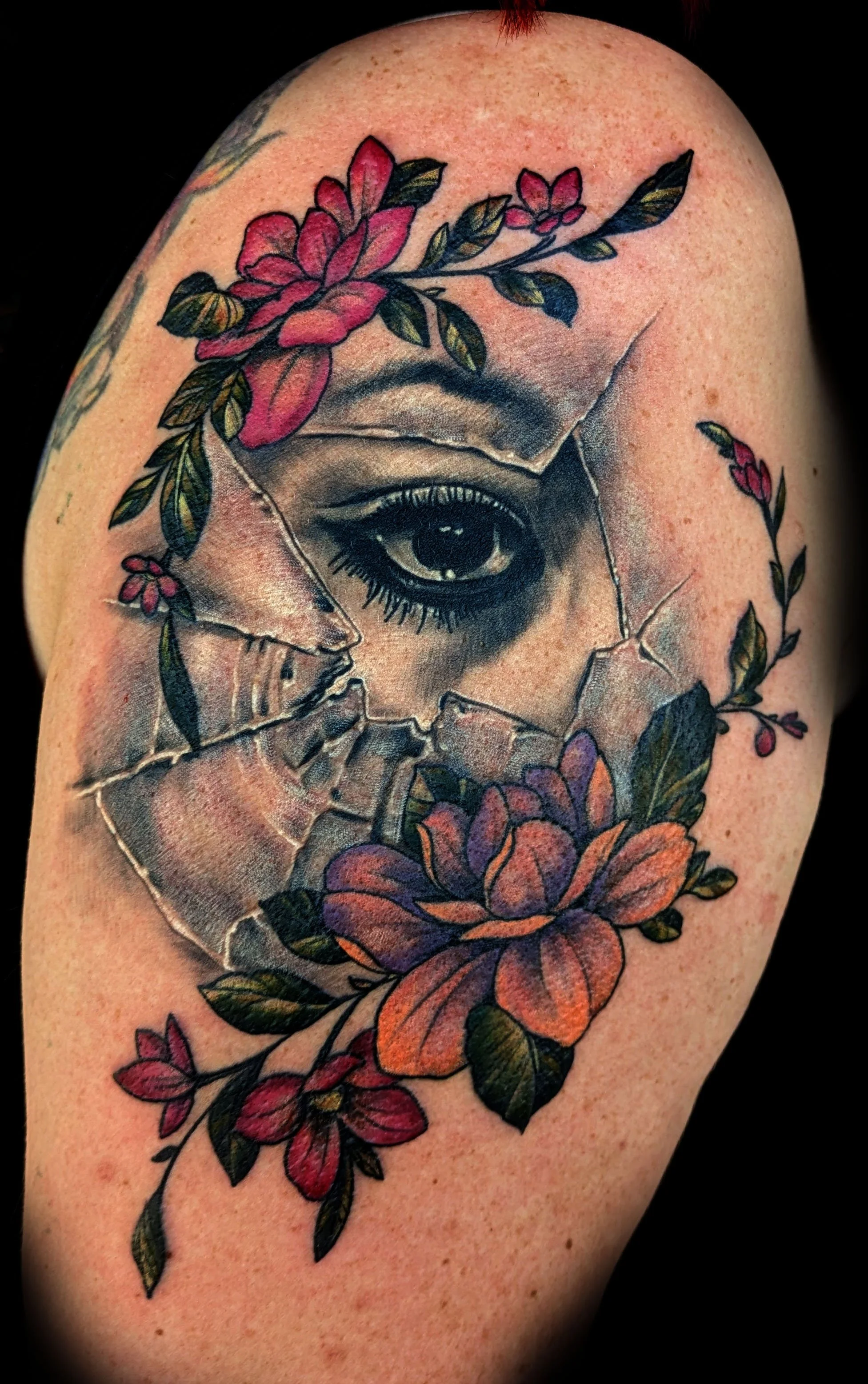 Tattoo of a realistic eye surrounded by colorful flowers and leaves, with some parts appearing as if the skin is cracked or broken.