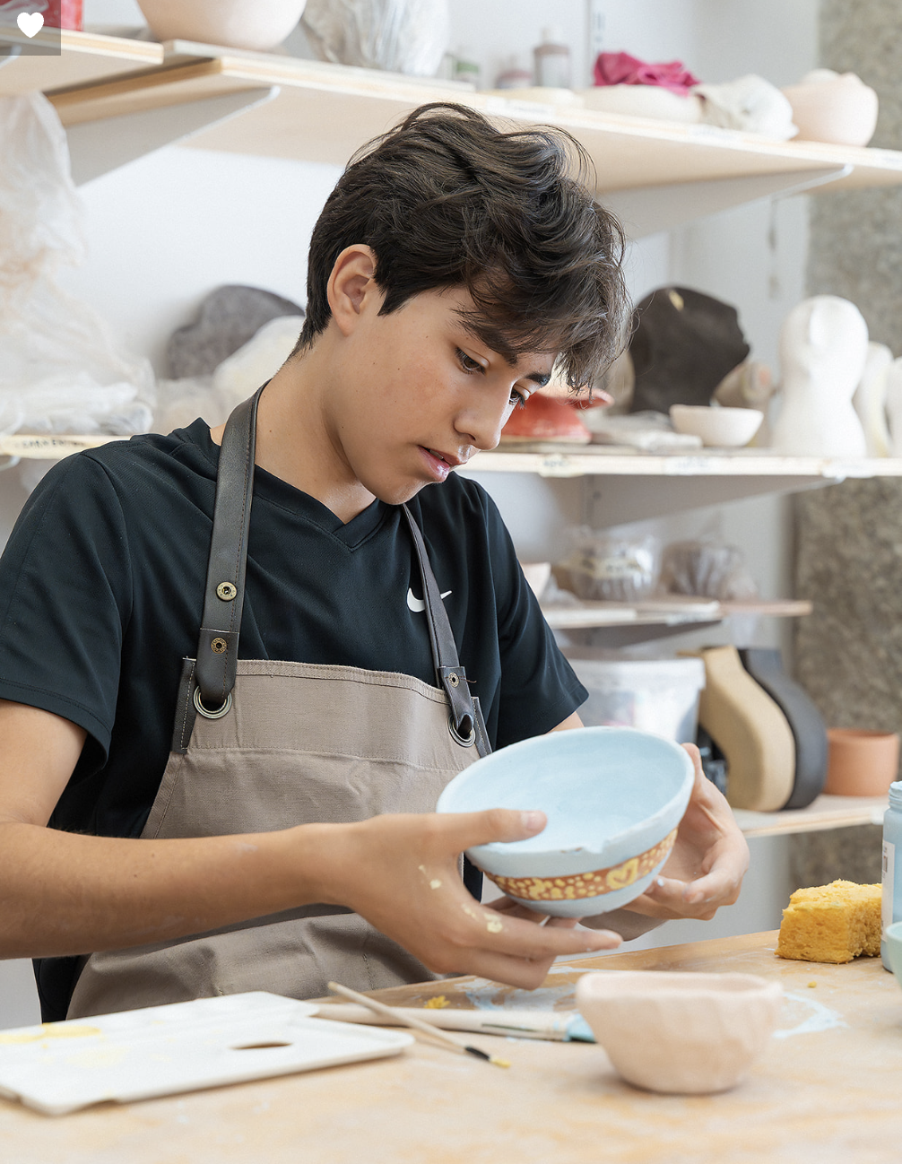 Young man working on pottery in a ceramics studio, holding a blue bowl and surrounded by pottery tools and finished pieces.