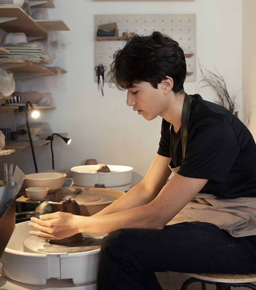 A young man is working on pottery at a pottery studio, shaping clay with his hands on a spinning wheel.