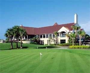 A scenic golf course with a putting green, a flag, and a large house in the background surrounded by palm trees under a clear blue sky.