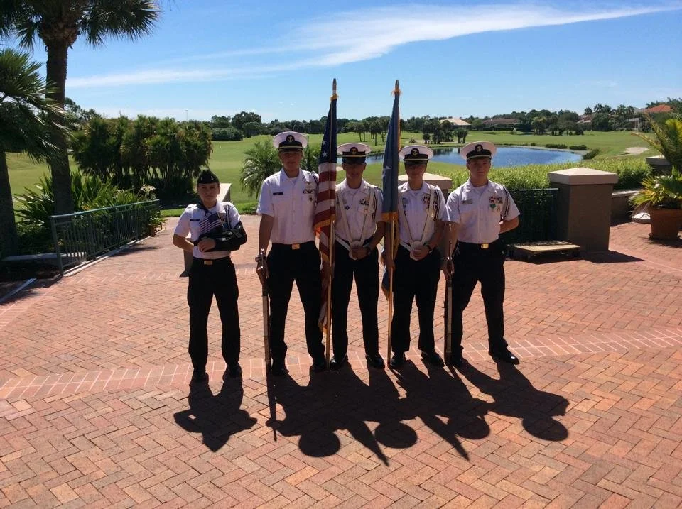 Five young individuals in uniform standing outdoors, holding flags, with a scenic view of a pond, greenery, and a golf course in the background on a sunny day.