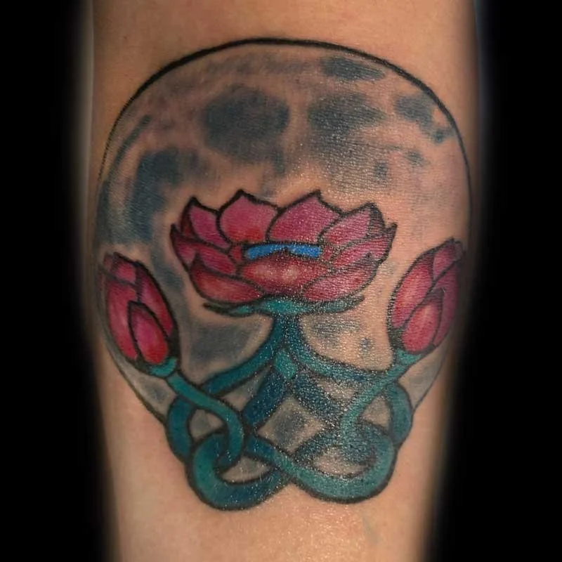 Tattoo of a pink lotus flower with blue-green stem inside a moon with cloud patterns.