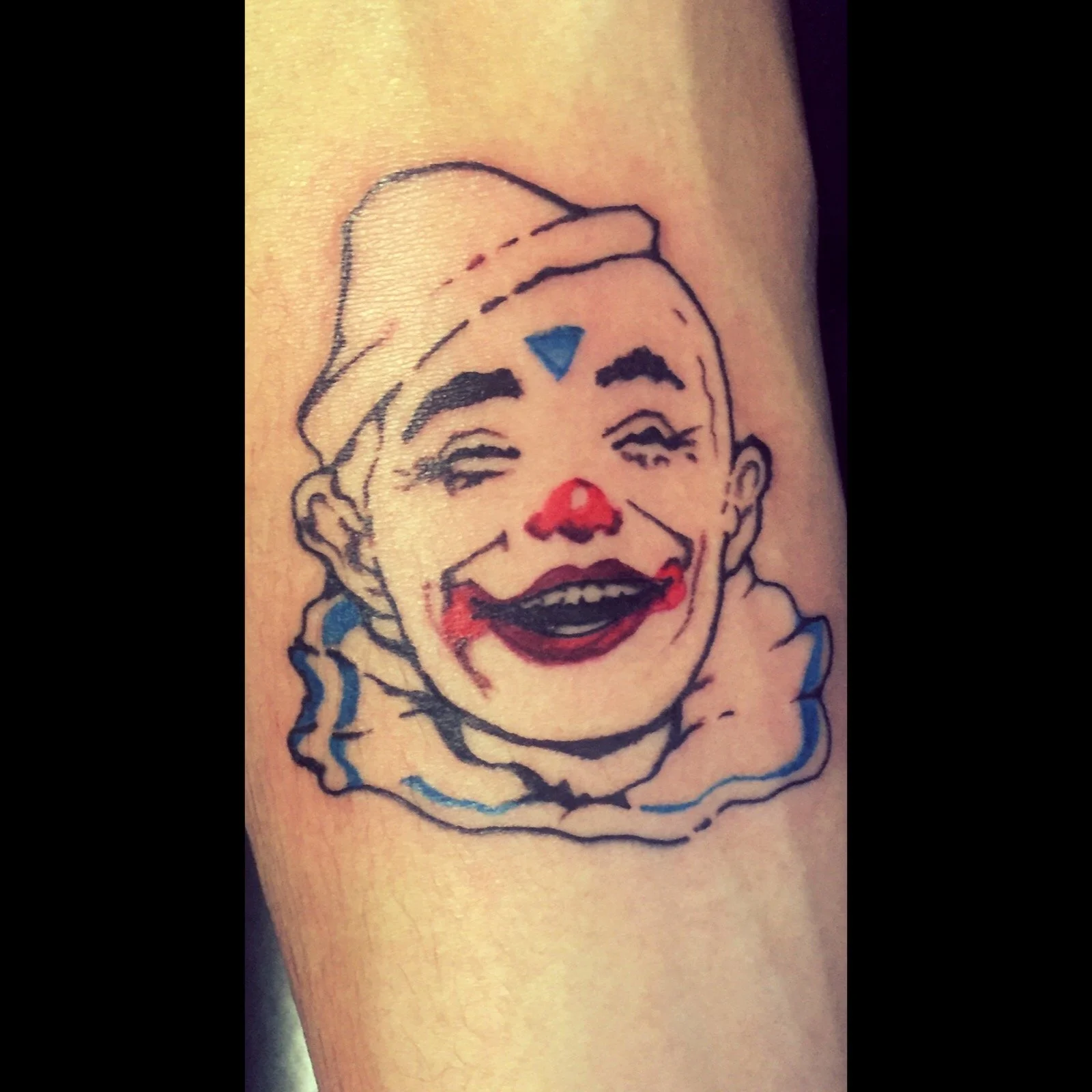 Tattoo of a clown face with a red nose, exaggerated smiling mouth, and makeup, wearing a hat, with a ruffled collar around the neck.