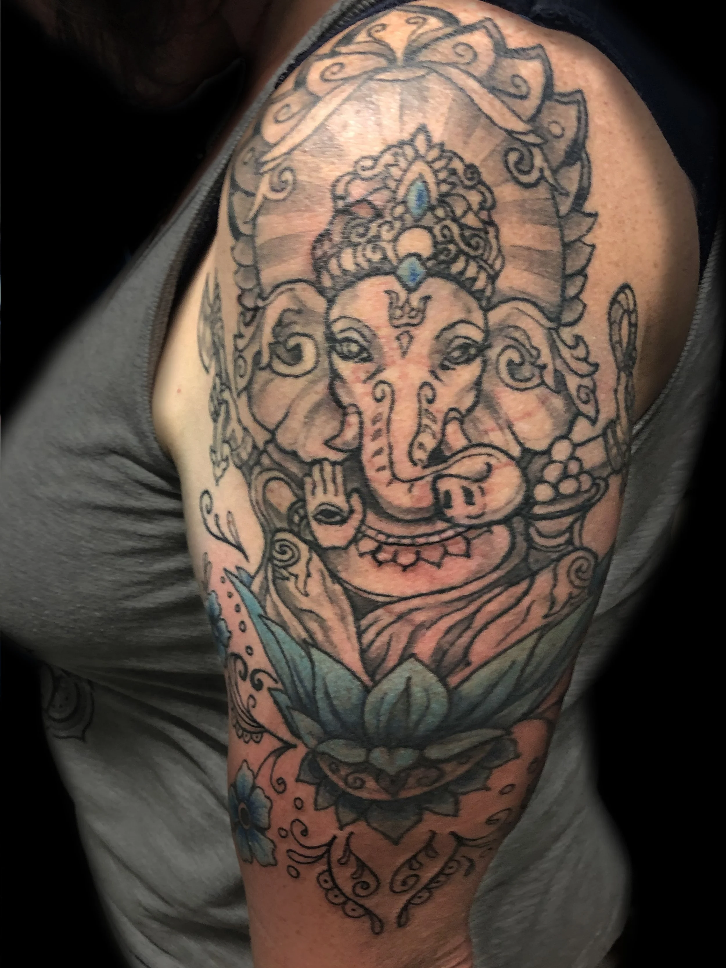 Tattoo of Ganesha on a person's upper arm, featuring a detailed depiction of the Hindu elephant-headed god with decorative elements and a blue lotus flower at the bottom.
