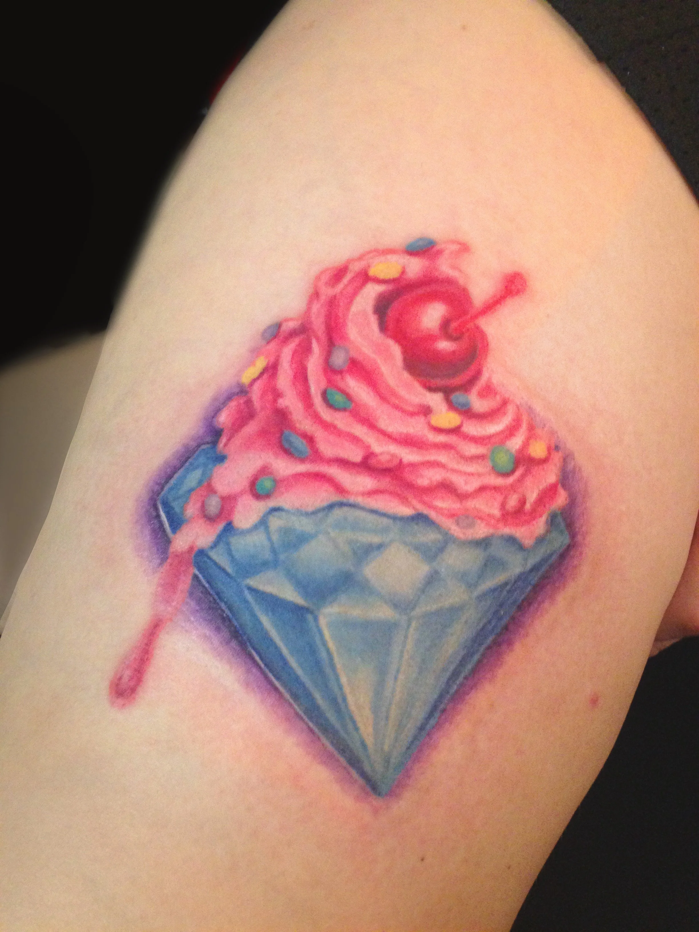 Tattoo of a cupcake with pink frosting, sprinkles, a cherry and a lit match, set on a blue diamond-shaped gemstone.