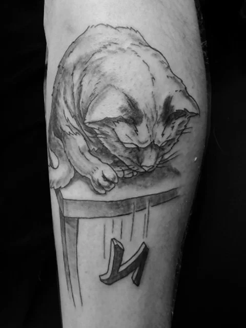 Black and gray tattoo of a sleeping cat curled up on a surface.