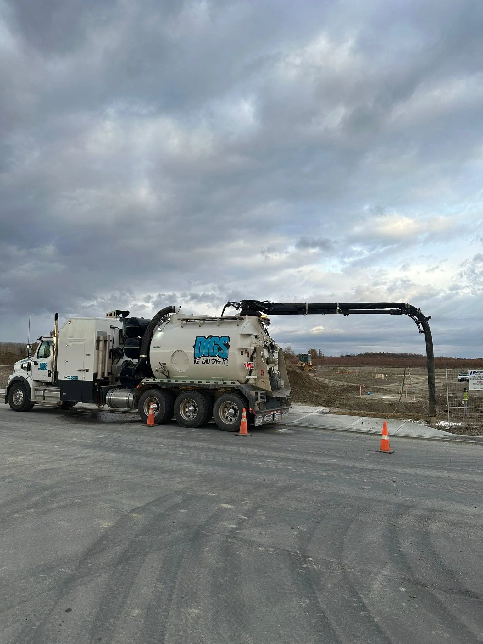 A vacuum truck with the logo 'Digs' and the slogan 'We Can Dig It!' is parked on a construction site, with an extendable hose reaching into the ground to perform excavation work. Orange traffic cones surround the truck, and a construction worker can be seen in the background under a cloudy sky.