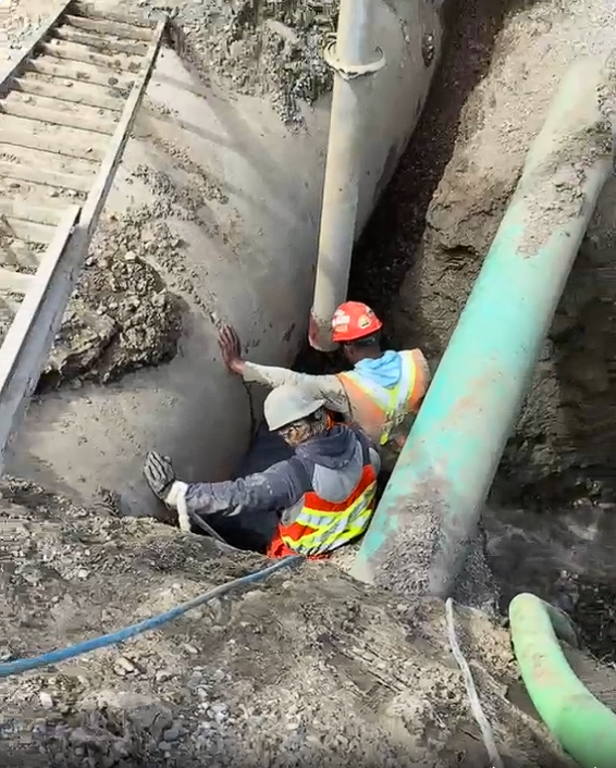 Two construction workers in safety gear are working in a trench with large pipes, one worker is using their hands to support the pipe while the other is assisting.