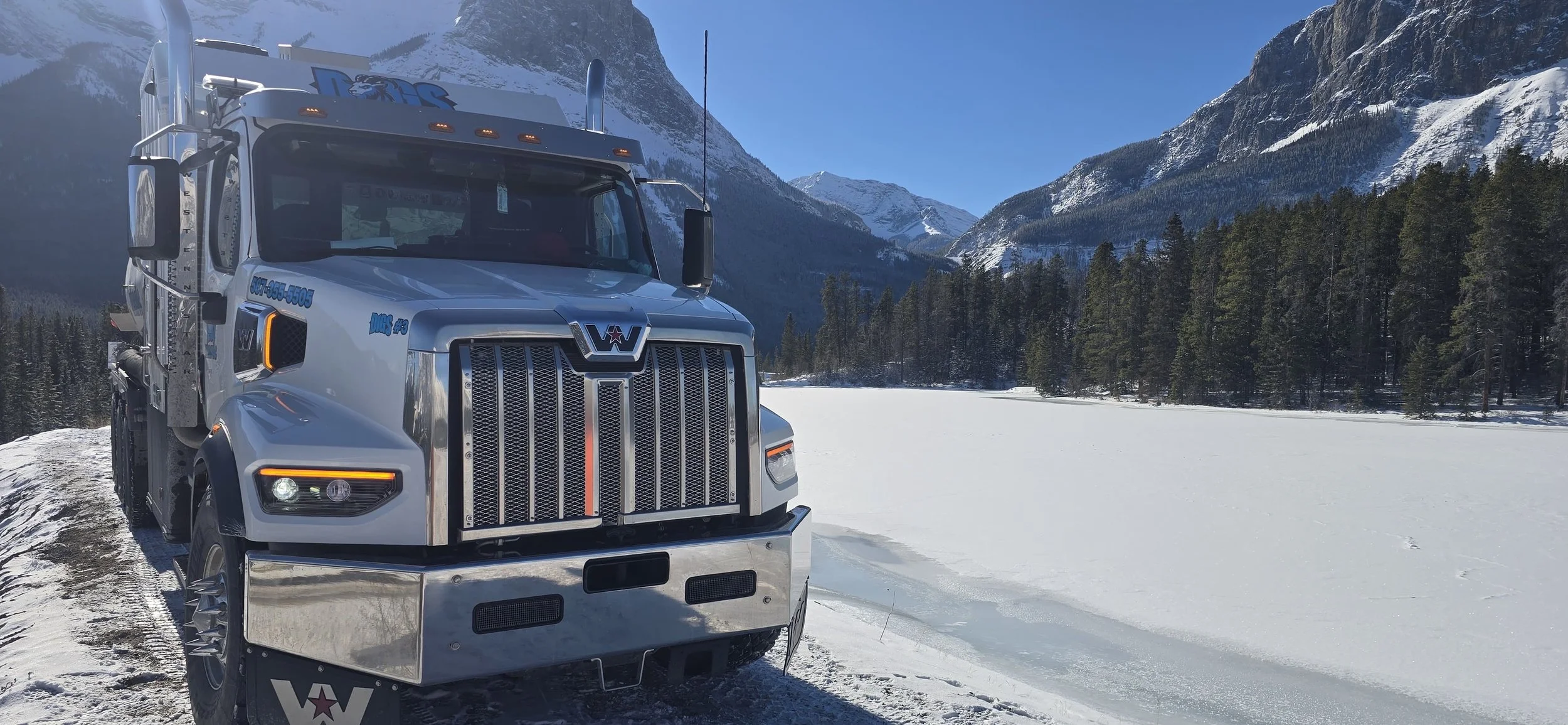 A large white semi-truck parked on a snowy landscape with a frozen lake, pine trees, and snow-covered mountains in the background under a clear blue sky.