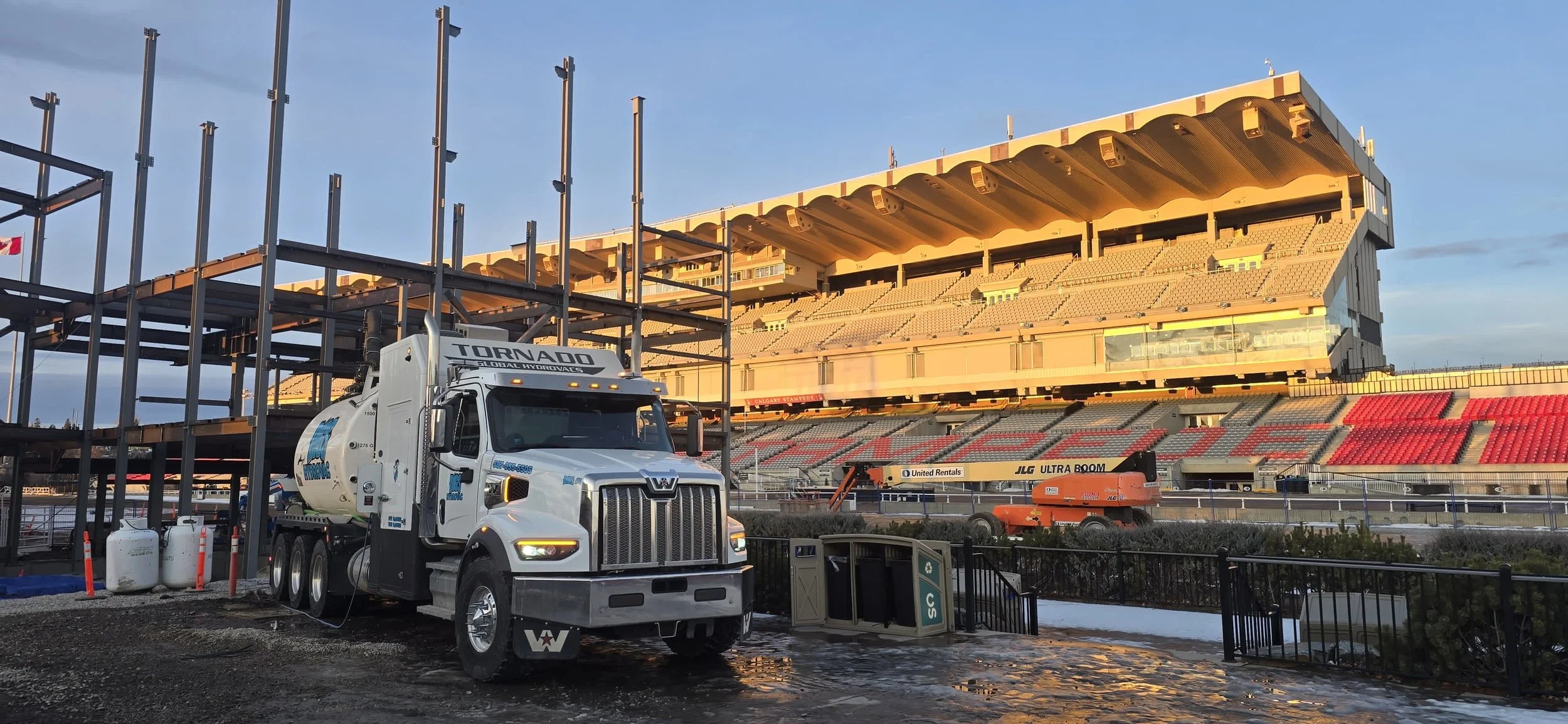Construction work at a stadium with a large white water tanker truck labeled 'TORNADO' and 'GLOBAL HYDROVAC' parked near scaffolding and a lift equipment, with empty seating in the background, during late afternoon or early evening.