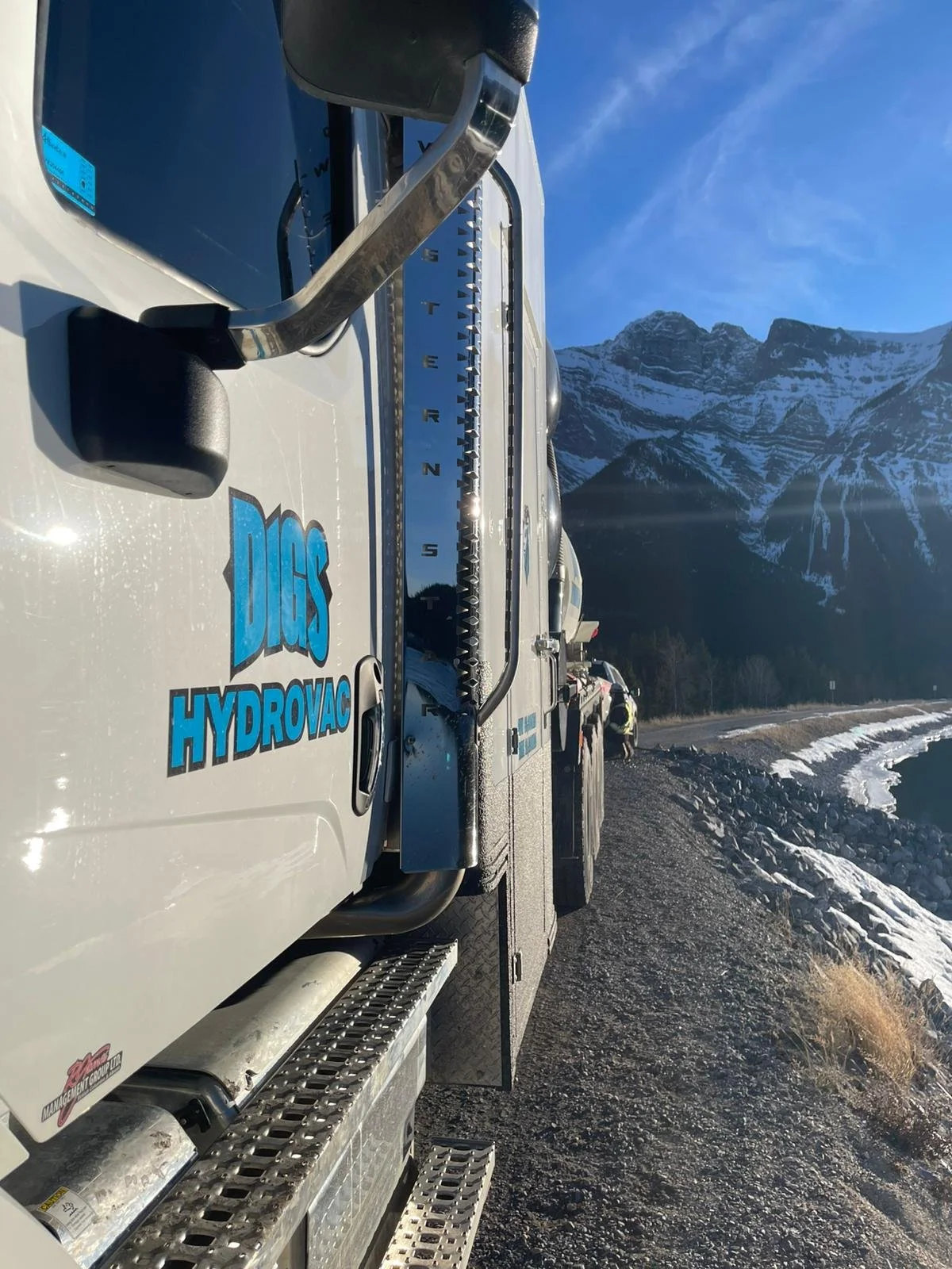 A white hydraulic service truck with the logo 'DGS Hydrovac' parked on a gravel road near a body of water, with snow-capped mountains in the background under a clear blue sky.