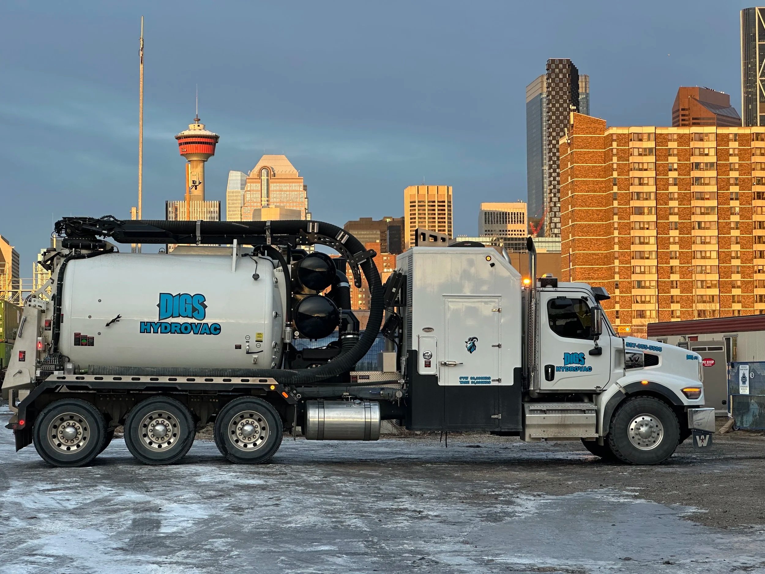 A large white vacuum truck labeled 'DIGS HYDROVAC' parked on icy ground with the city skyline of Calgary in the background.