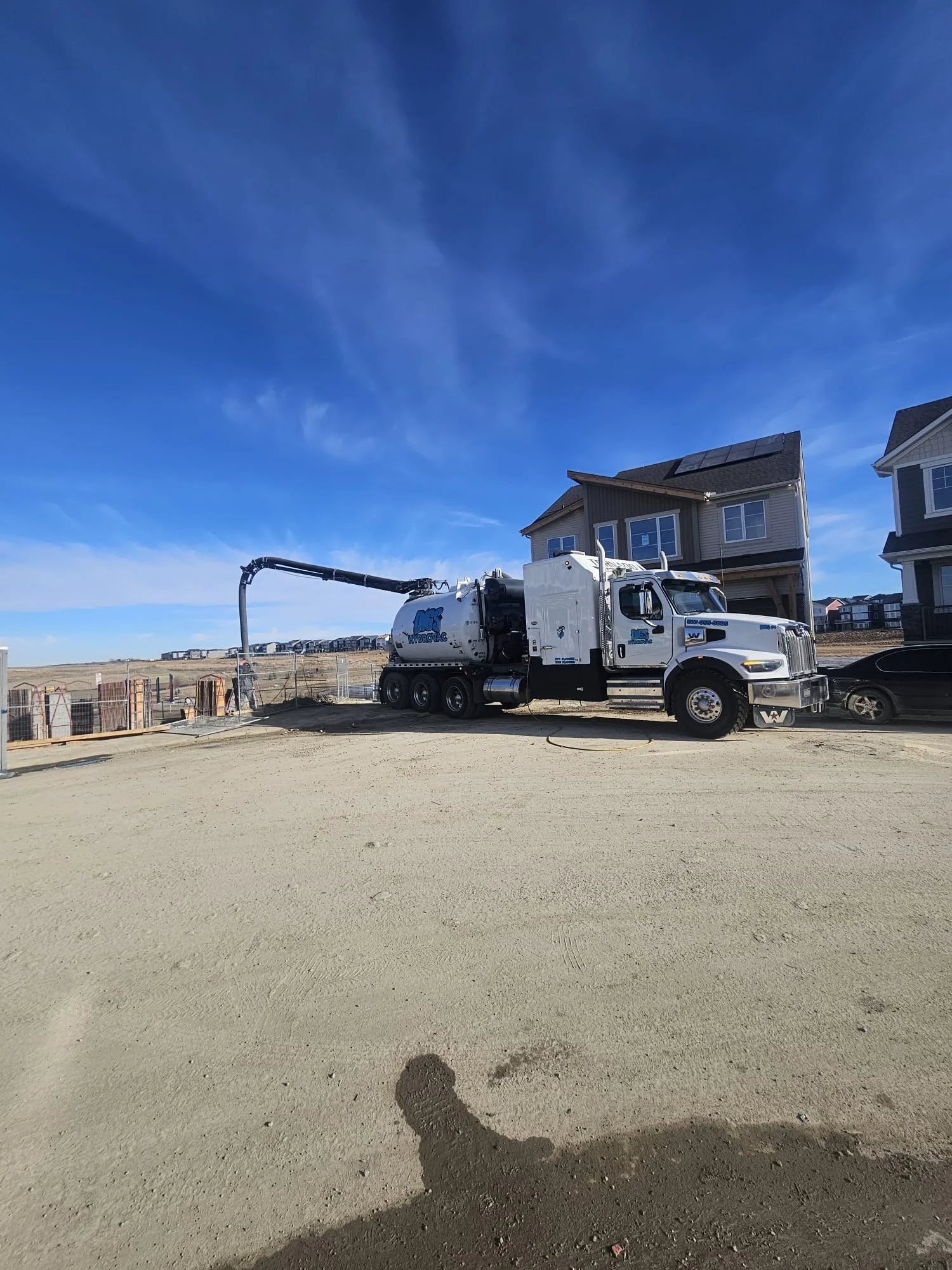 A white vacuum truck with a long hose attached is on a construction site, with new houses in the background and a partly cloudy blue sky overhead.