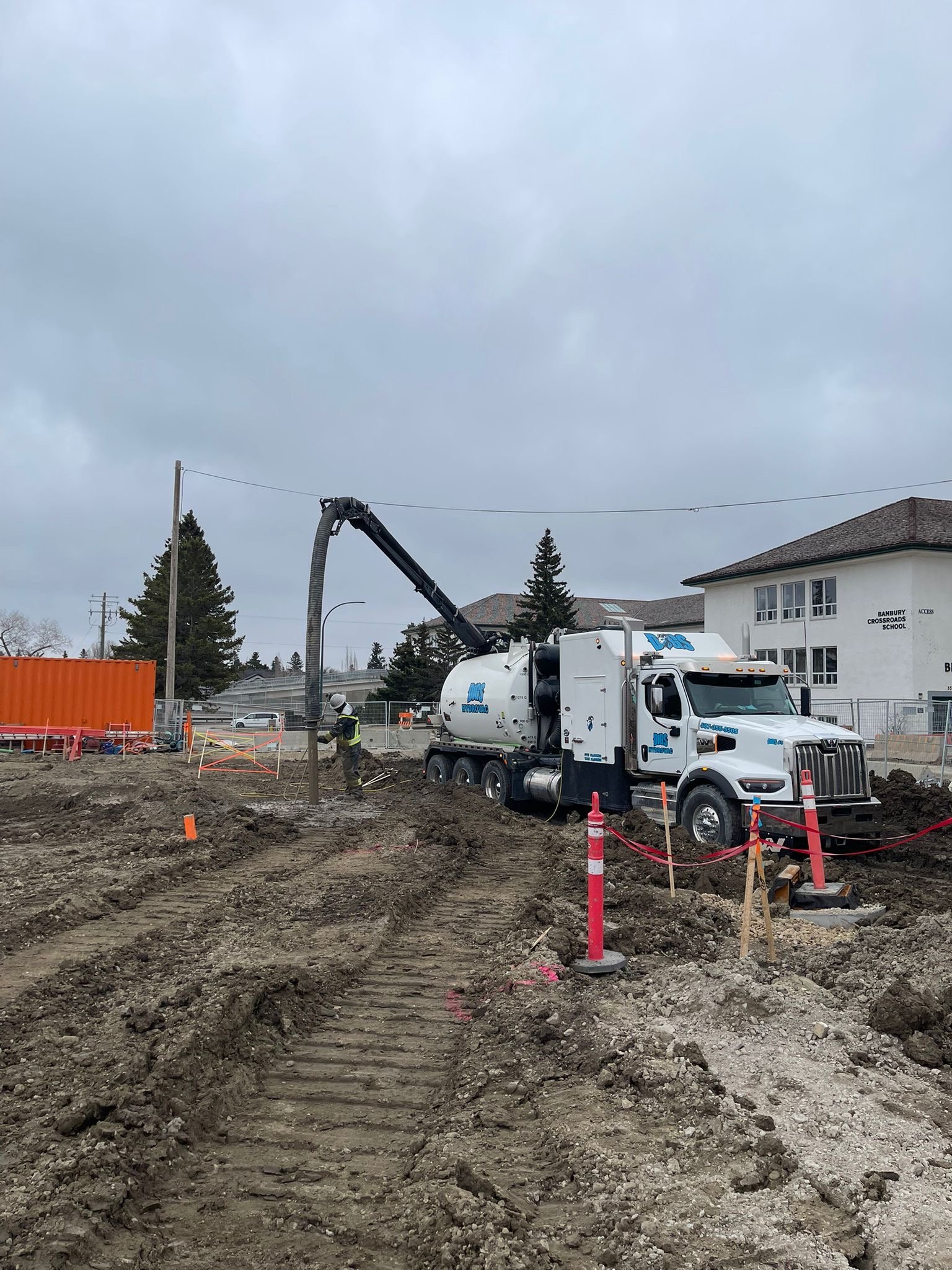 Construction site with a large white cement mixer truck and workers digging in the dirt. Overcast sky, nearby buildings, trees, and construction barriers are visible.