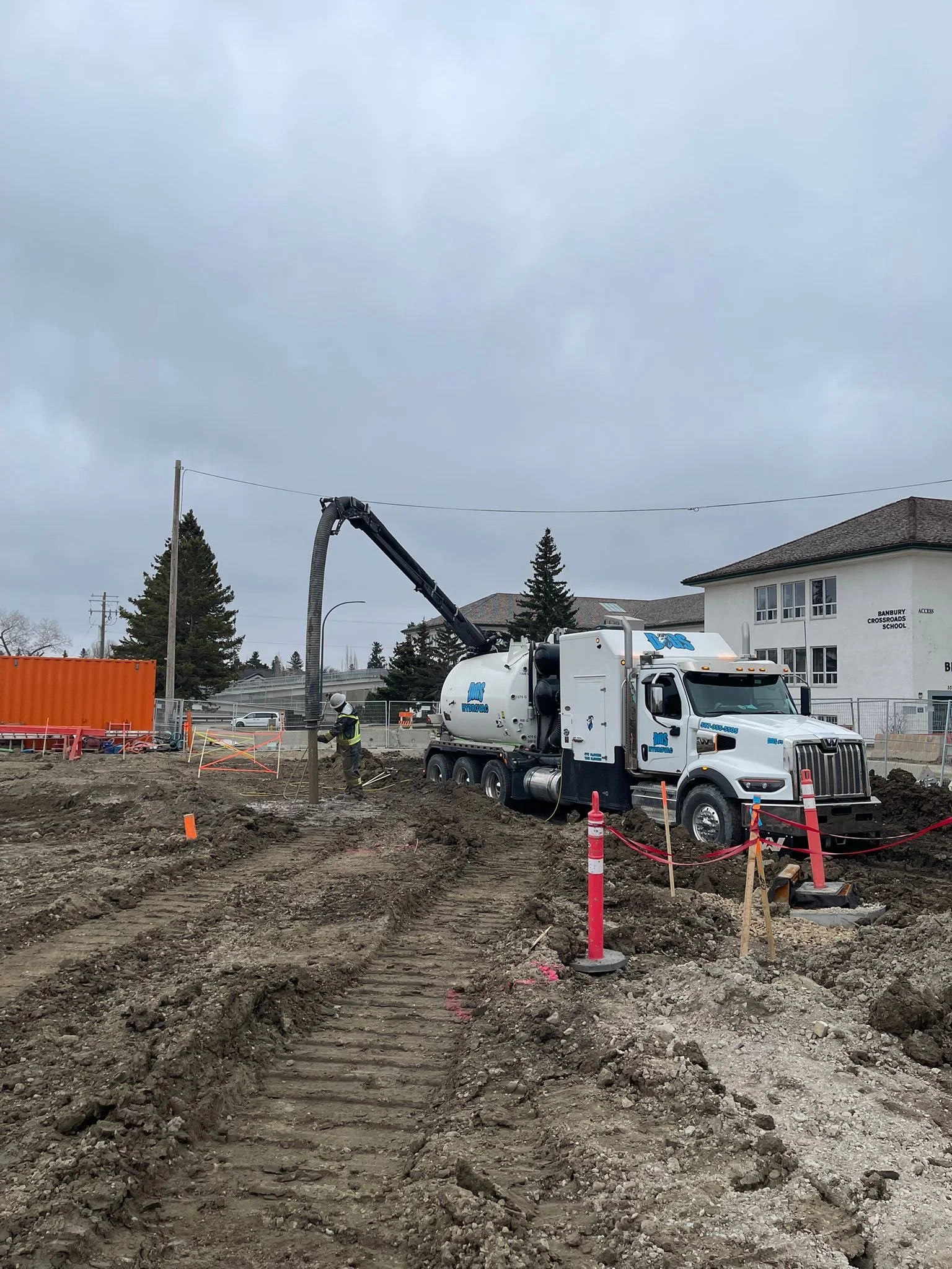 Construction site with a large white cement mixer truck and workers digging in the dirt. Overcast sky, nearby buildings, trees, and construction barriers are visible.