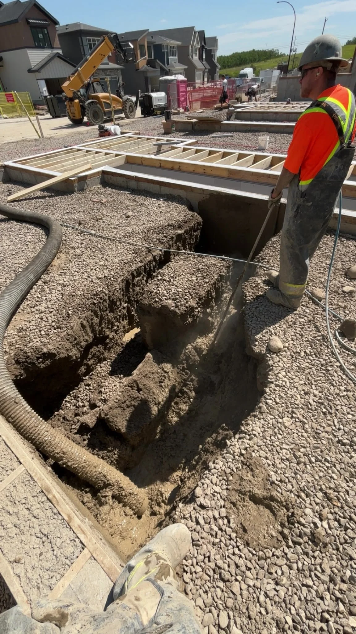 DIGS Hydrovac worker in safety gear at a large excavation site with wooden foundation framing, equipment, and nearby buildings under construction.