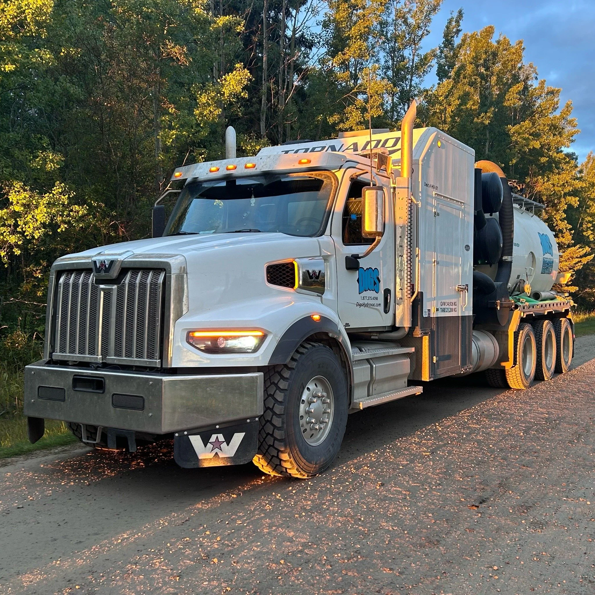 A white septic service truck parked on the side of a dirt road with trees in the background, illuminated by sunlight.