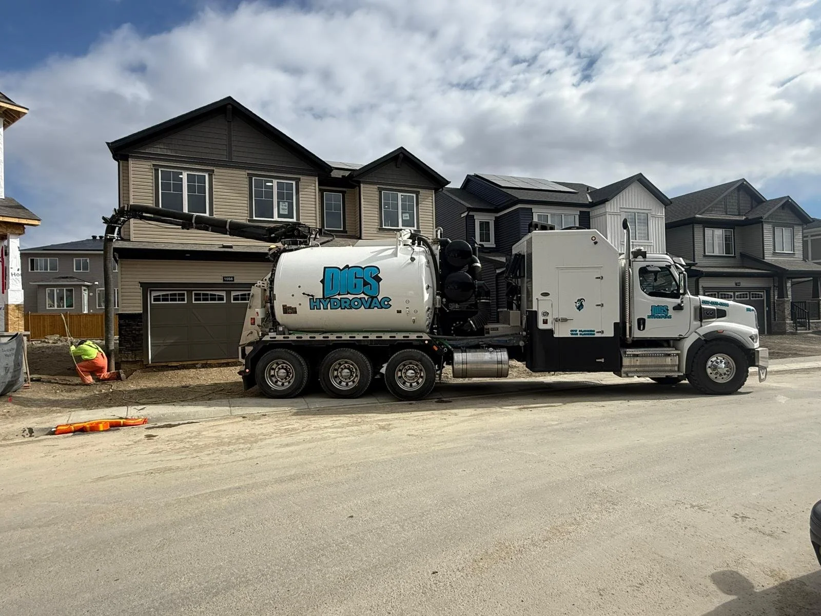 A construction worker in a yellow safety vest and orange pants working on a landscaping project in front of a modern multi-story house with a garage, while a large white vacuum truck with the words 'Digs HydroVac' is parked on the street.