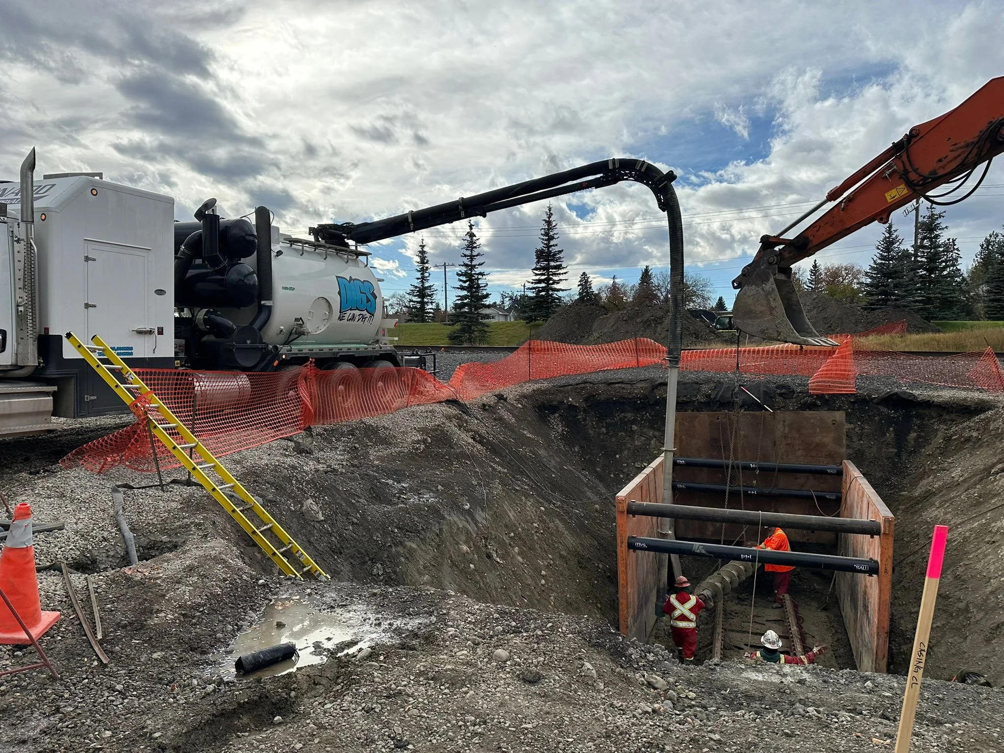 Construction workers in safety gear working in a large underground excavation with orange safety barriers and construction equipment, including a vacuum truck with a large hose and an excavator.