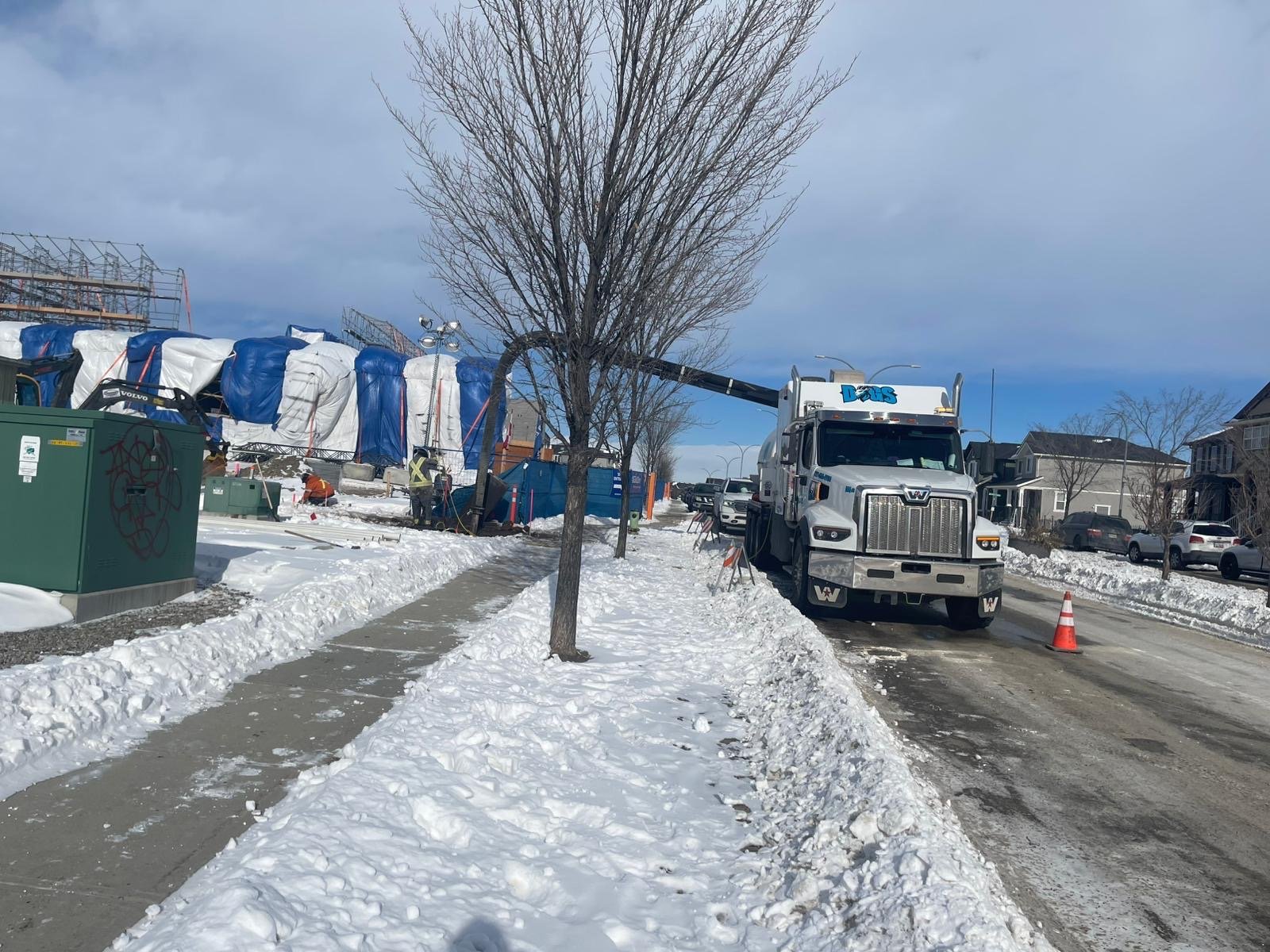 Street with snow and ice, a tree without leaves, a utility truck parked on the side, and construction in the background covered with tarps.