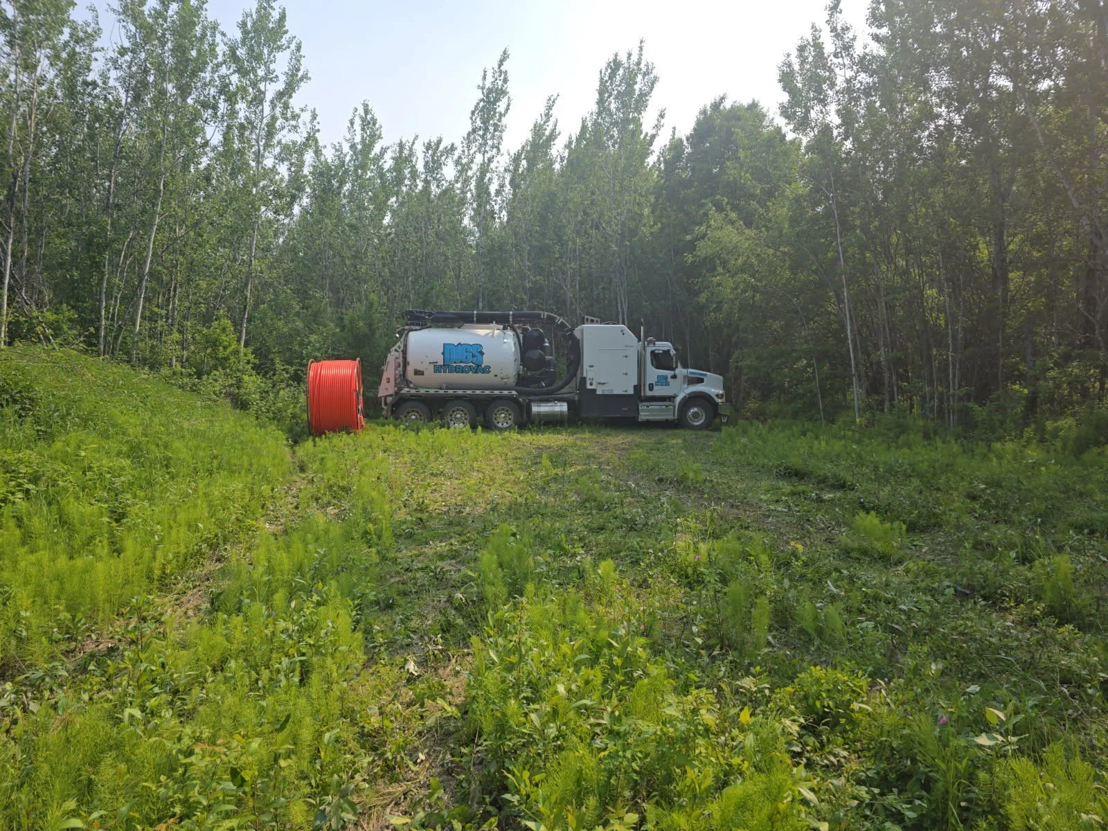A hydro Excavation truck with the logo 'DIGS Hydrovac' on its side is parked on a clearing in a wooded area. There is a large orange spool of tubing on the ground nearby and trees surrounding the area under a bright, clear sky.