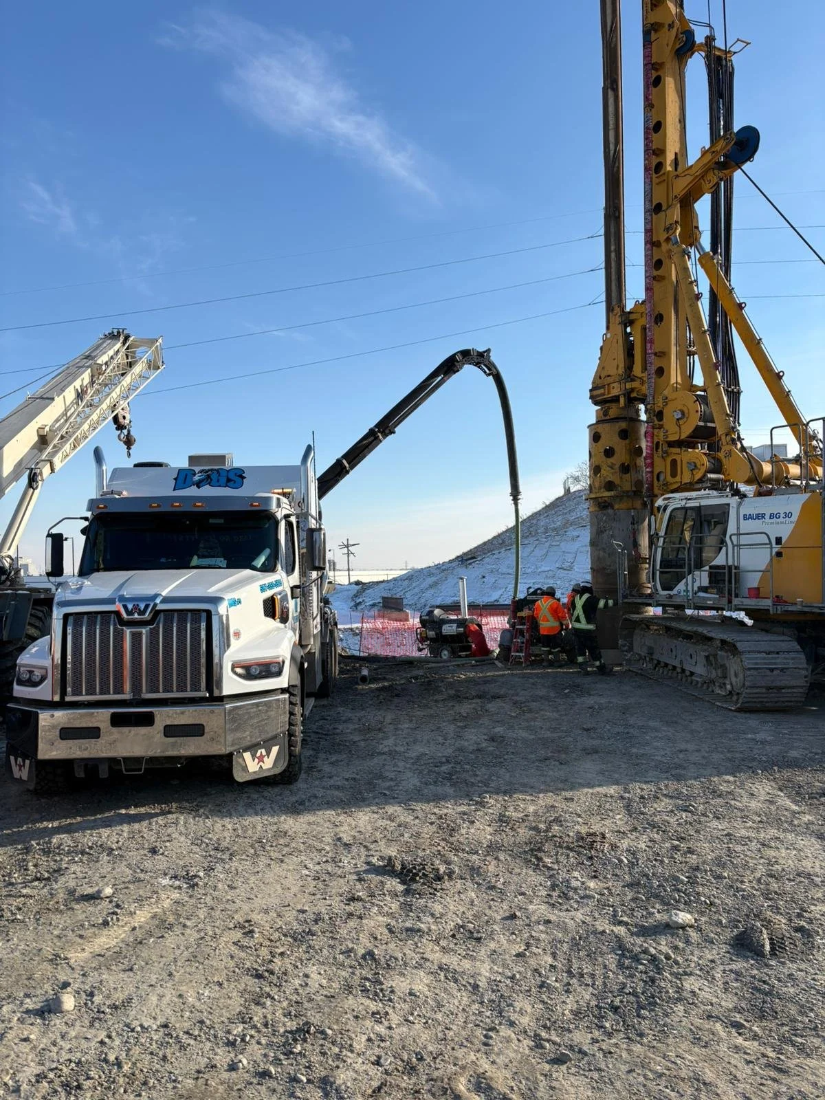Construction site with a white truck and a yellow drilling machine, two workers in safety vests are working beside a small portable generator, snow on the hill in the background, clear blue sky.