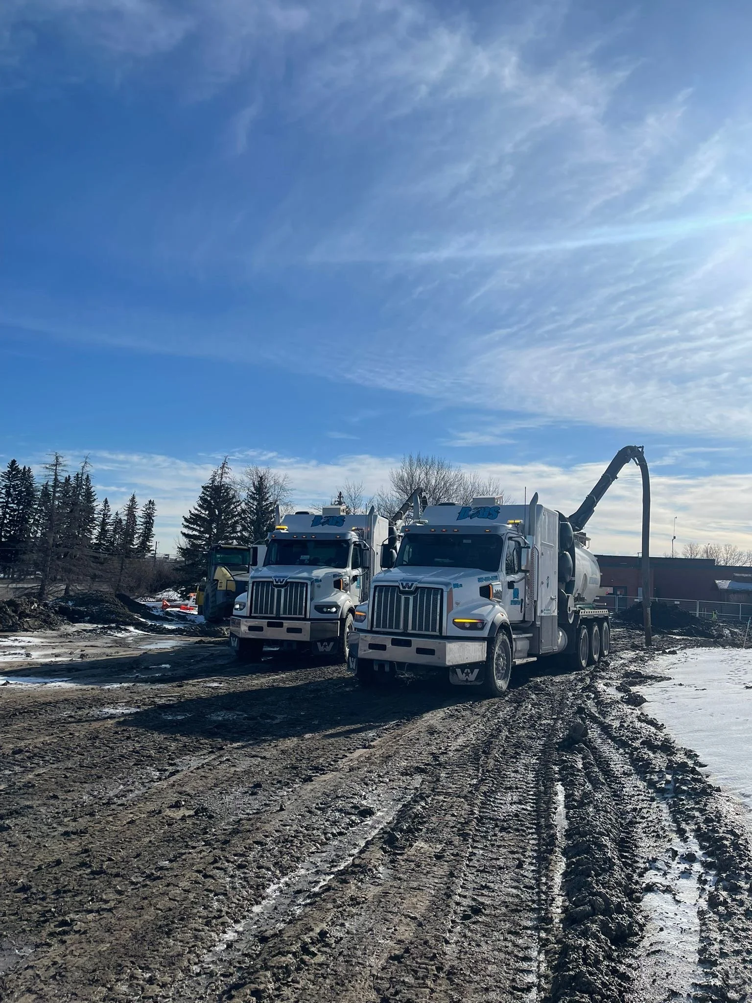 Two large white dump trucks with the word 'Deros' on top, parked on a muddy construction site with tire tracks, snow, leafless trees, and a partly cloudy blue sky.