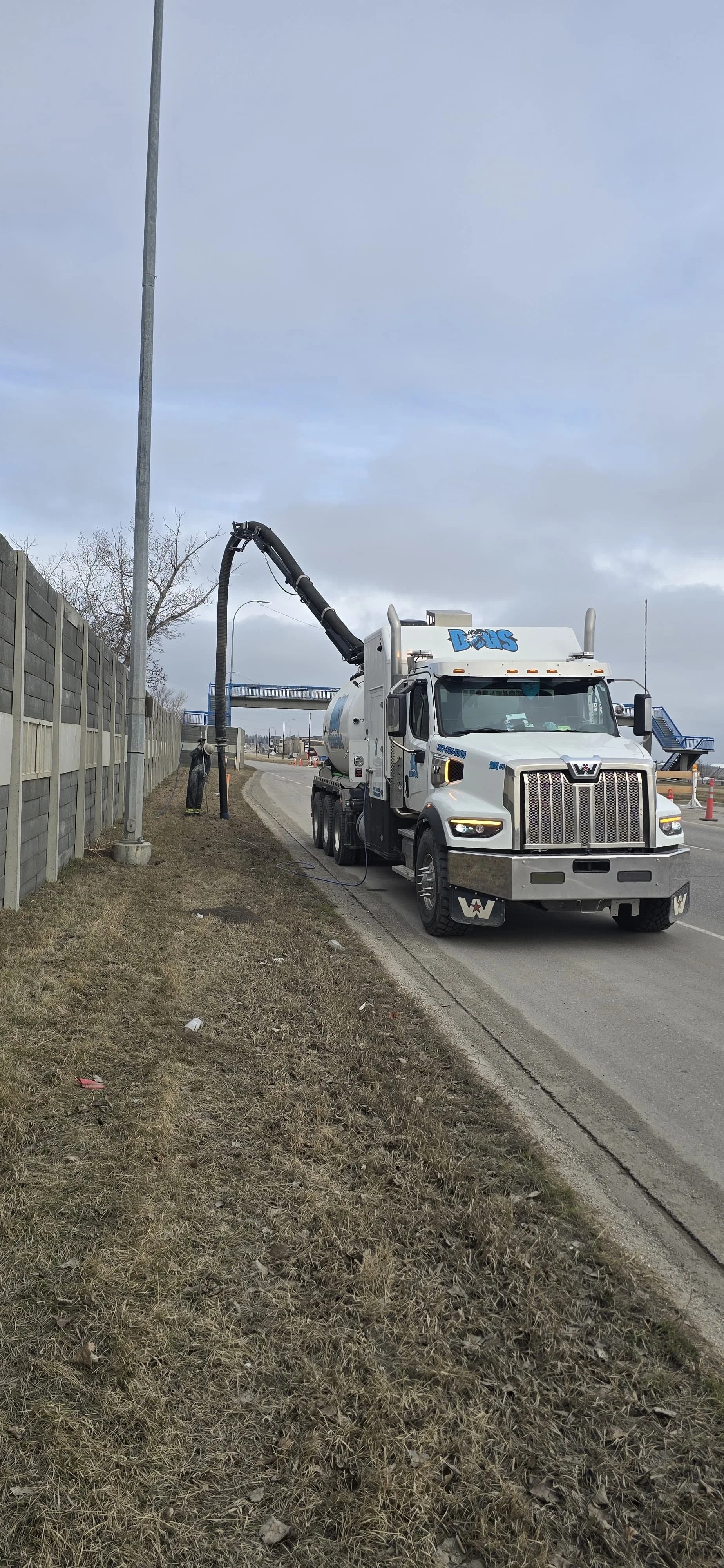 A street scene with a large white vacuum truck with the logo 'Dogs' on top, suctioning dirt or debris from the ground along the sidewalk, with a tall fence on the left and a pedestrian overpass in the background.