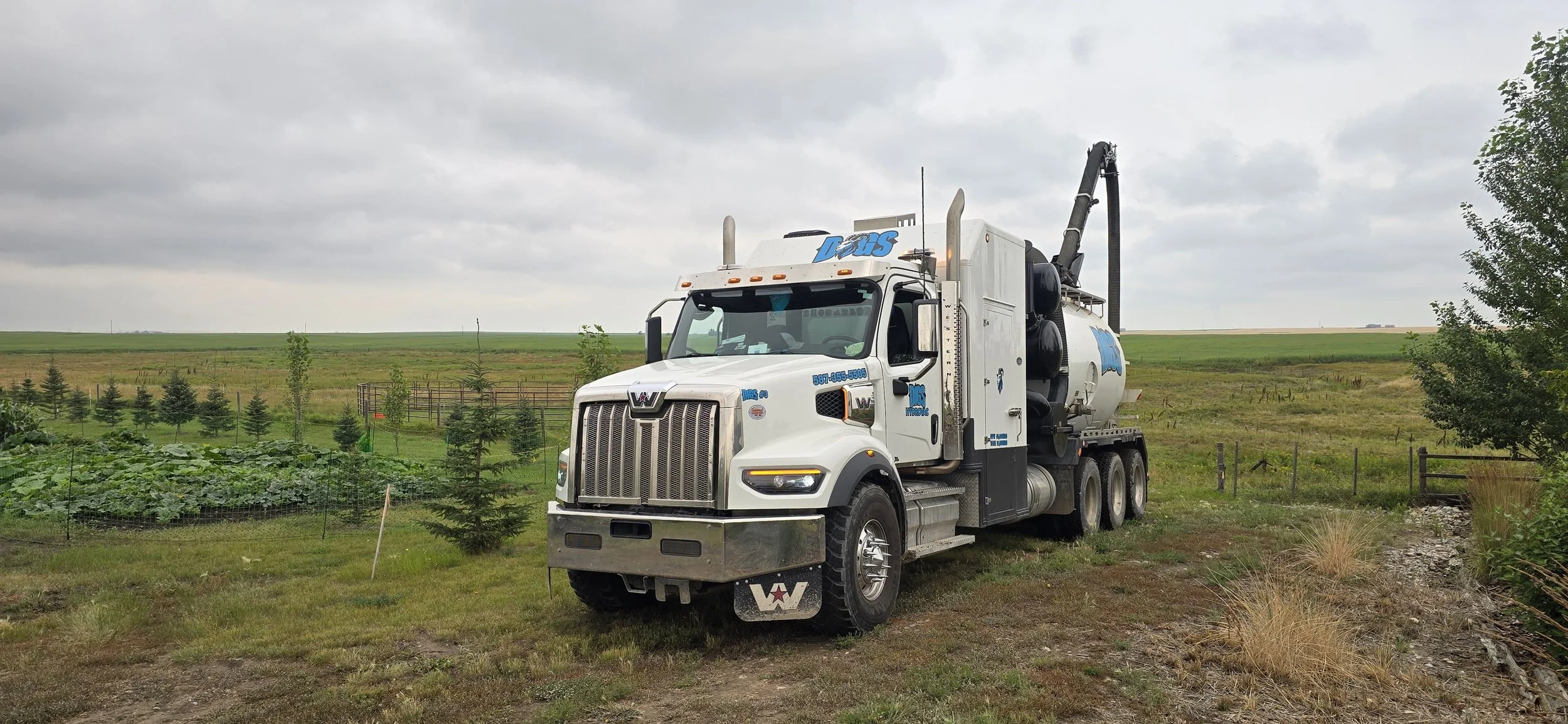 White DIGS Hydrovac vacuum truck parked on a grassy field with trees and plants around, under a cloudy sky.