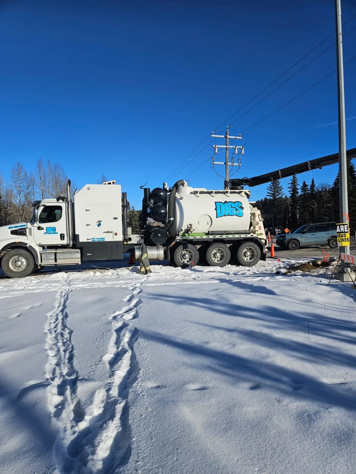 A large white utility truck with the word 'DIGS' on the side, parked on snow-covered ground near utility poles and power lines.