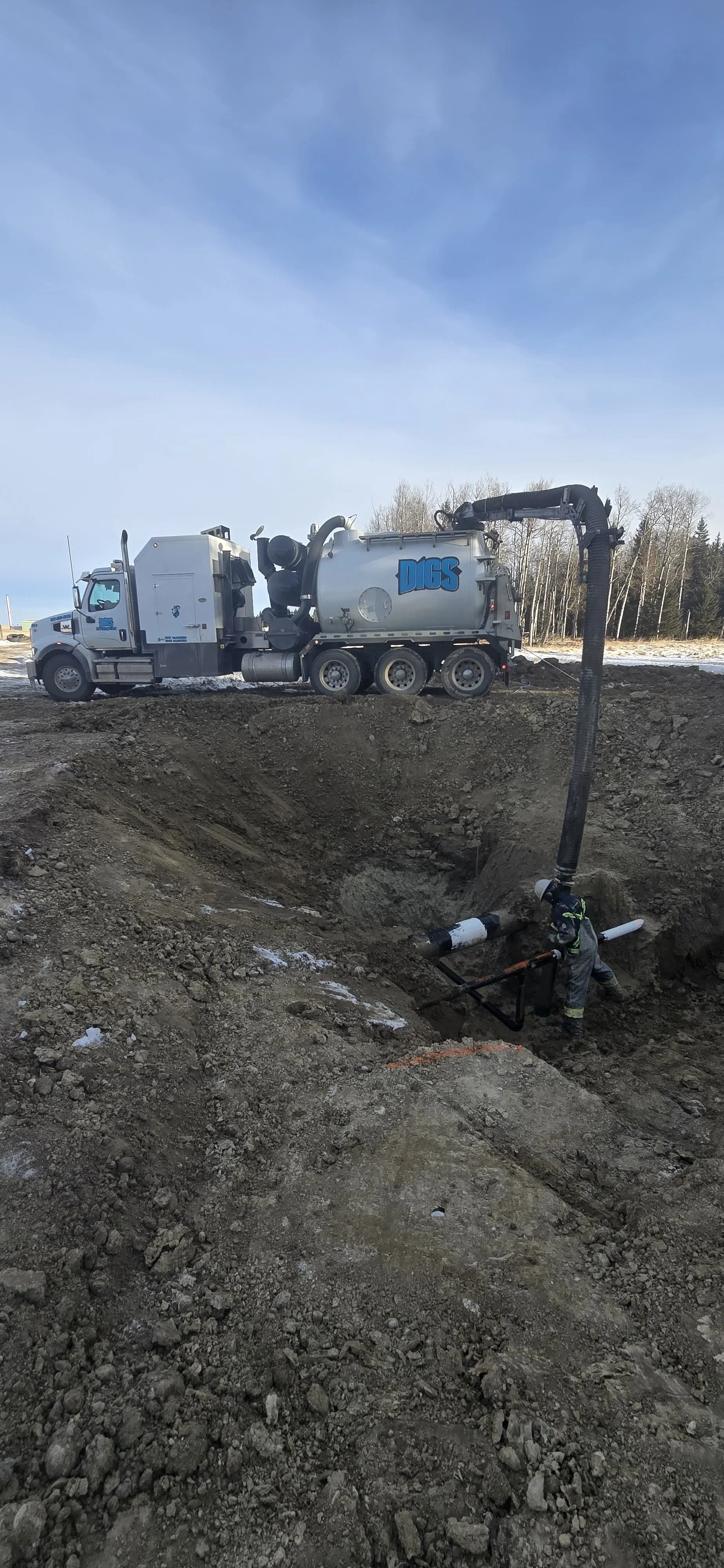 A construction worker operating a large vacuum excavation truck labeled 'DIGS' in a dirt excavation site on a clear day with a blue sky and sparse trees in the background.