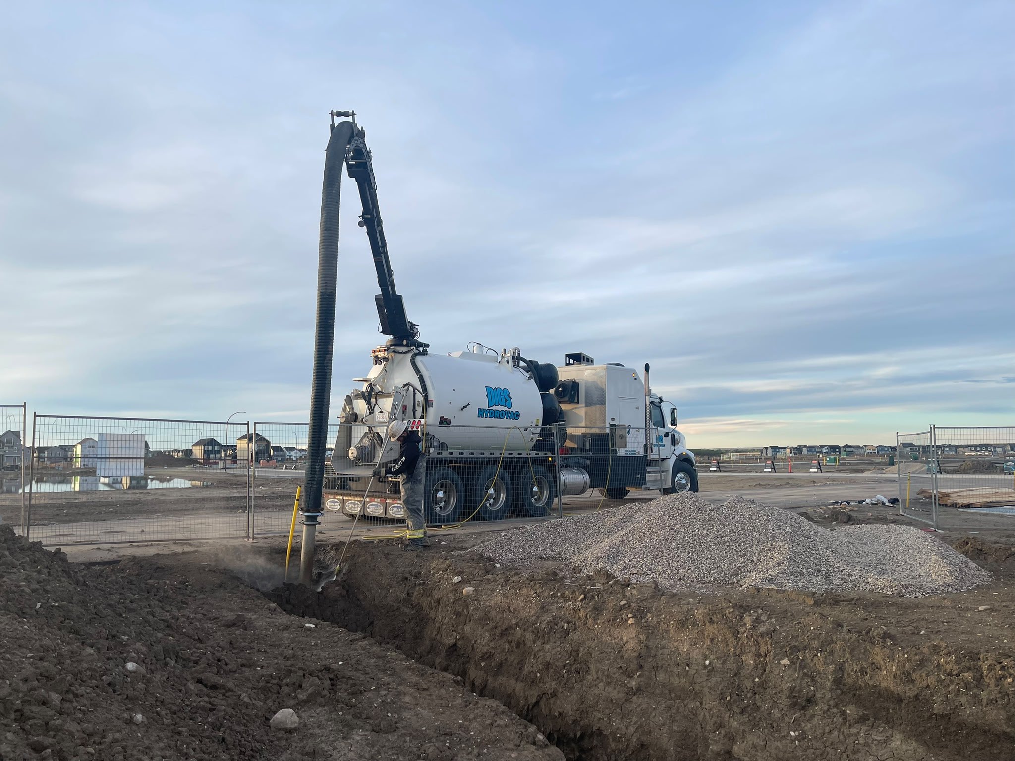 Construction workers and a large vacuum truck on a construction site, with a deep trench in the foreground and residential houses in the background.