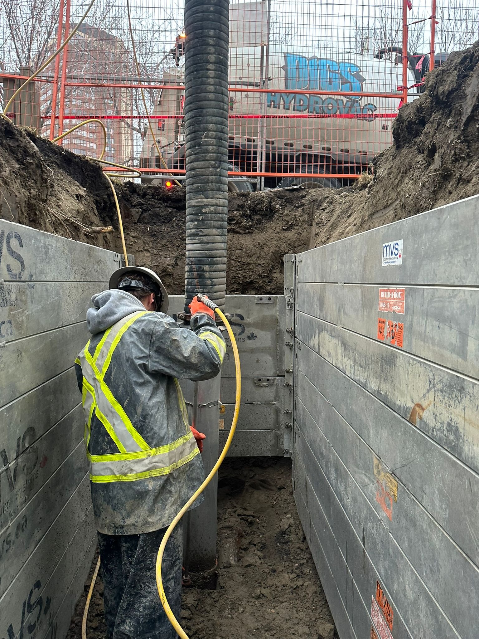 Construction worker in safety gear working in a trench with steel walls, excavation equipment, and a giant black pipe.