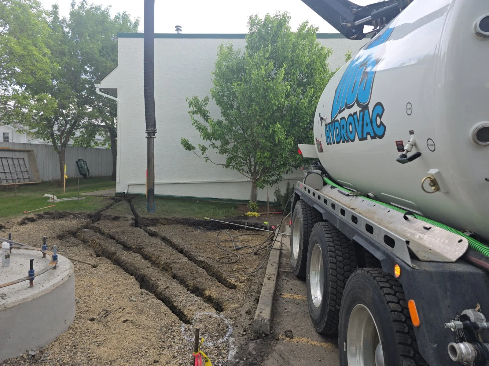 Construction site with a concrete truck on the right and freshly excavated soil with trenches and marking sticks in the foreground, a small white building with a tree in the background.