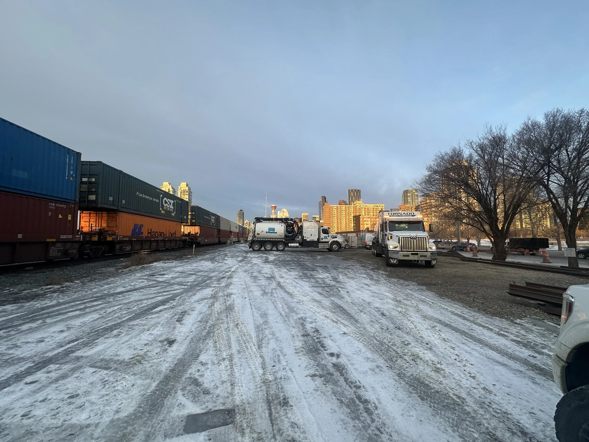 A snowy industrial area with train cars on the left, trucks and equipment in the center, and city buildings in the background, under a cloudy sky.