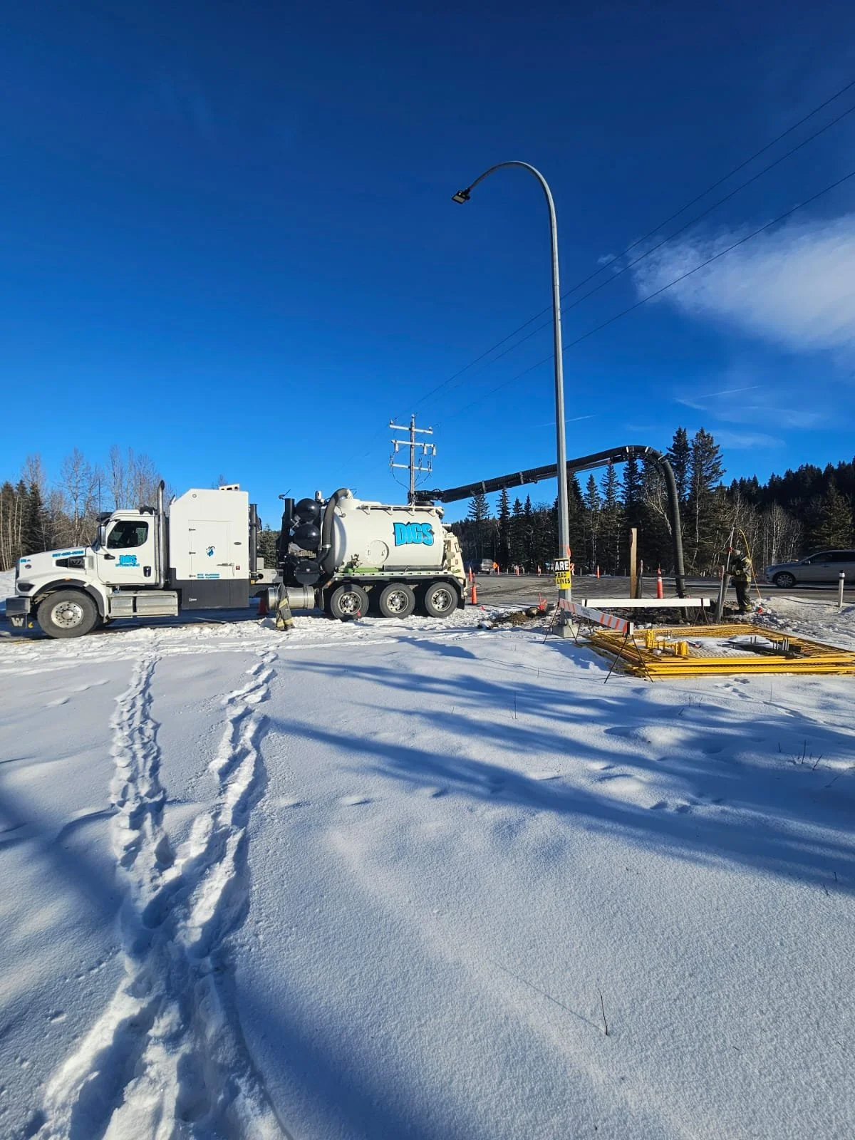 Water utility truck with long hose in snow-covered area, blue sky and trees in the background.