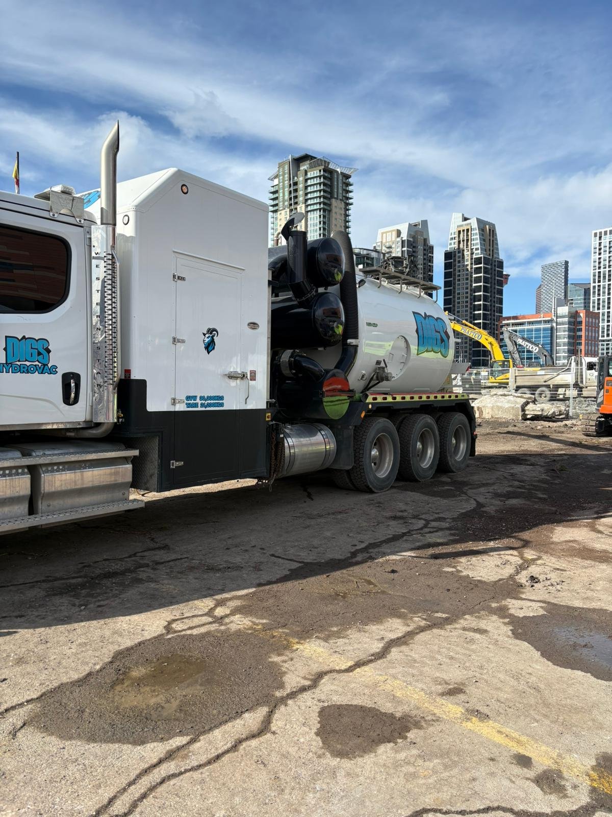 Construction site with a white water tanker truck labeled 'Digs Hydrovac' and machinery, with a city skyline in the background.