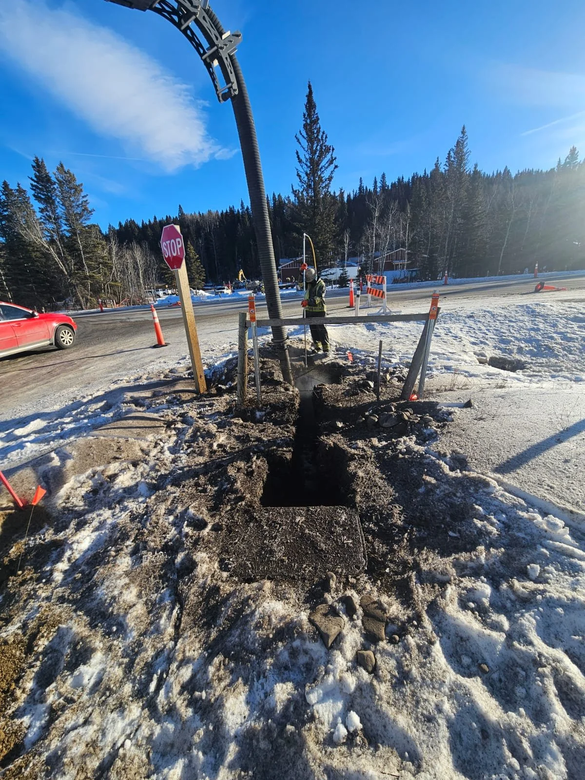 Workers installing a utility pole on the side of a snowy road, with traffic cones and a stop sign nearby, in a rural area with trees and mountains in the background.