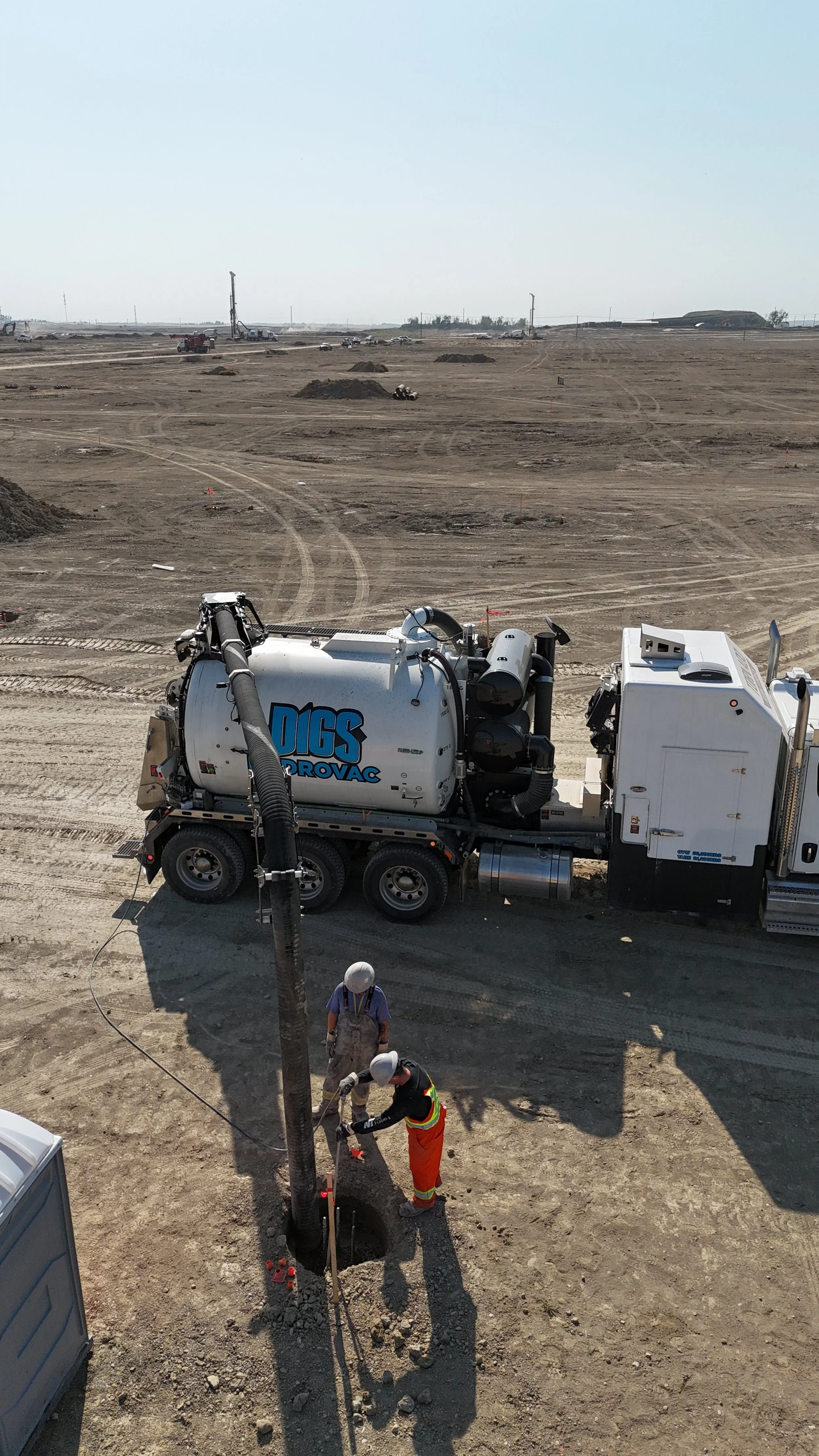 Construction workers in safety gear working on underground utility installation at a large construction site. There is a vacuum truck labeled Digs Provac alongside them.